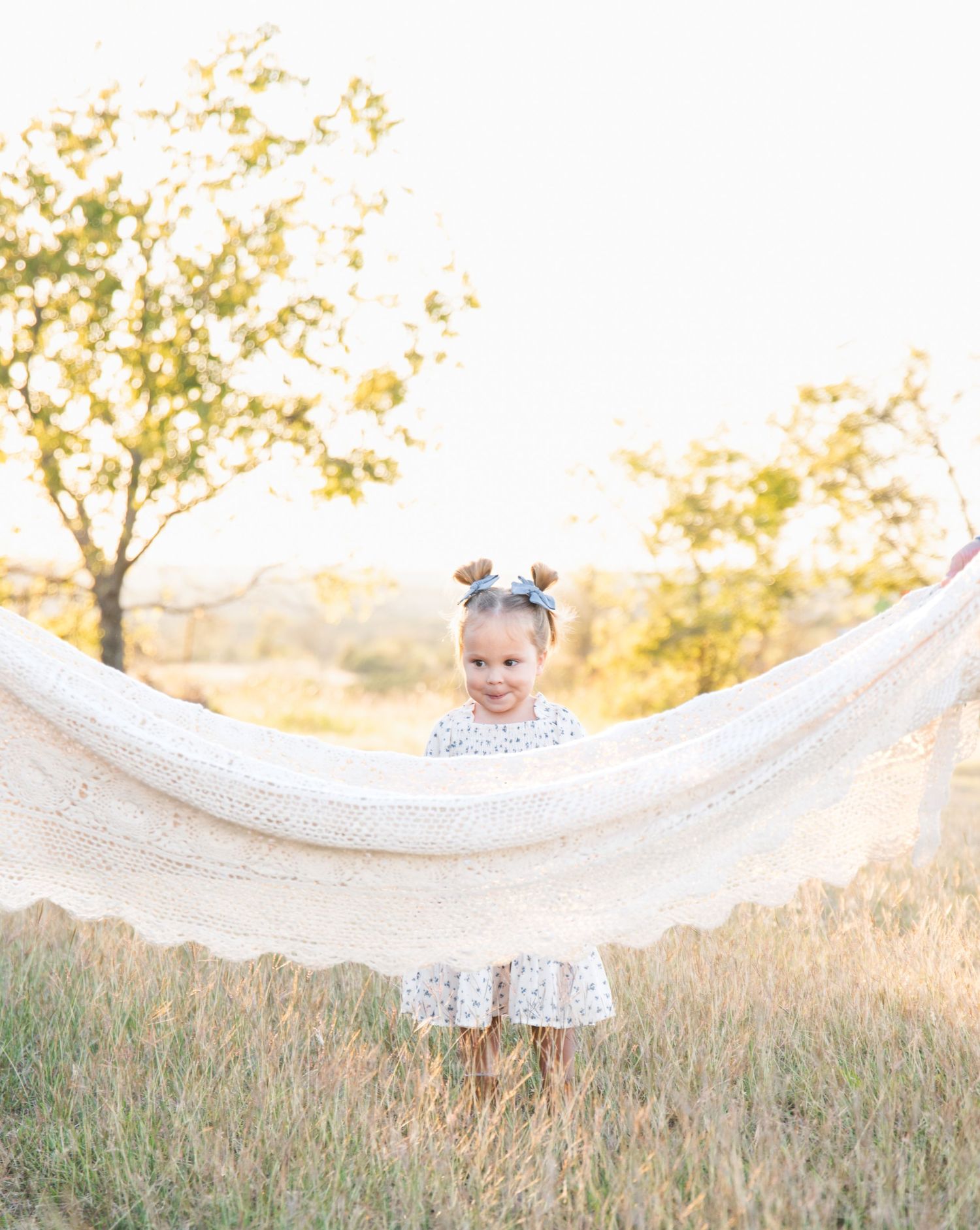 A small child in a polka dot dress holds a white fabric in a sun-lit field.