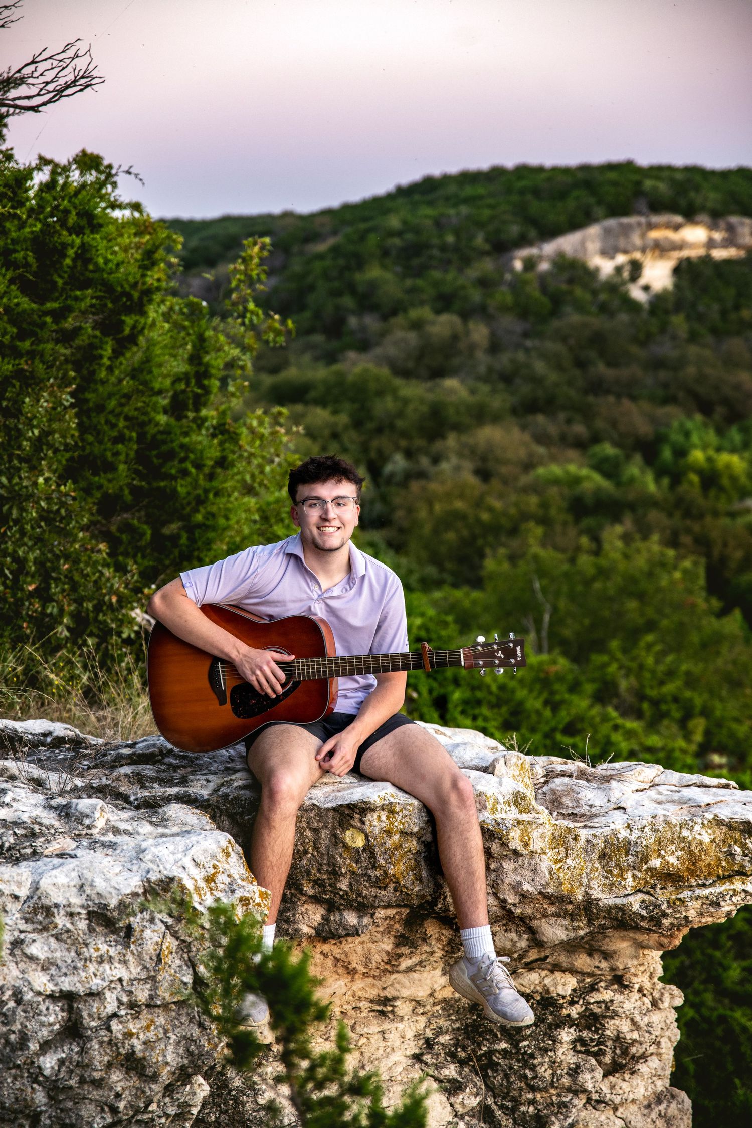 Acoustic guitar player sitting on limestone cliff edge with hill country landscape background.