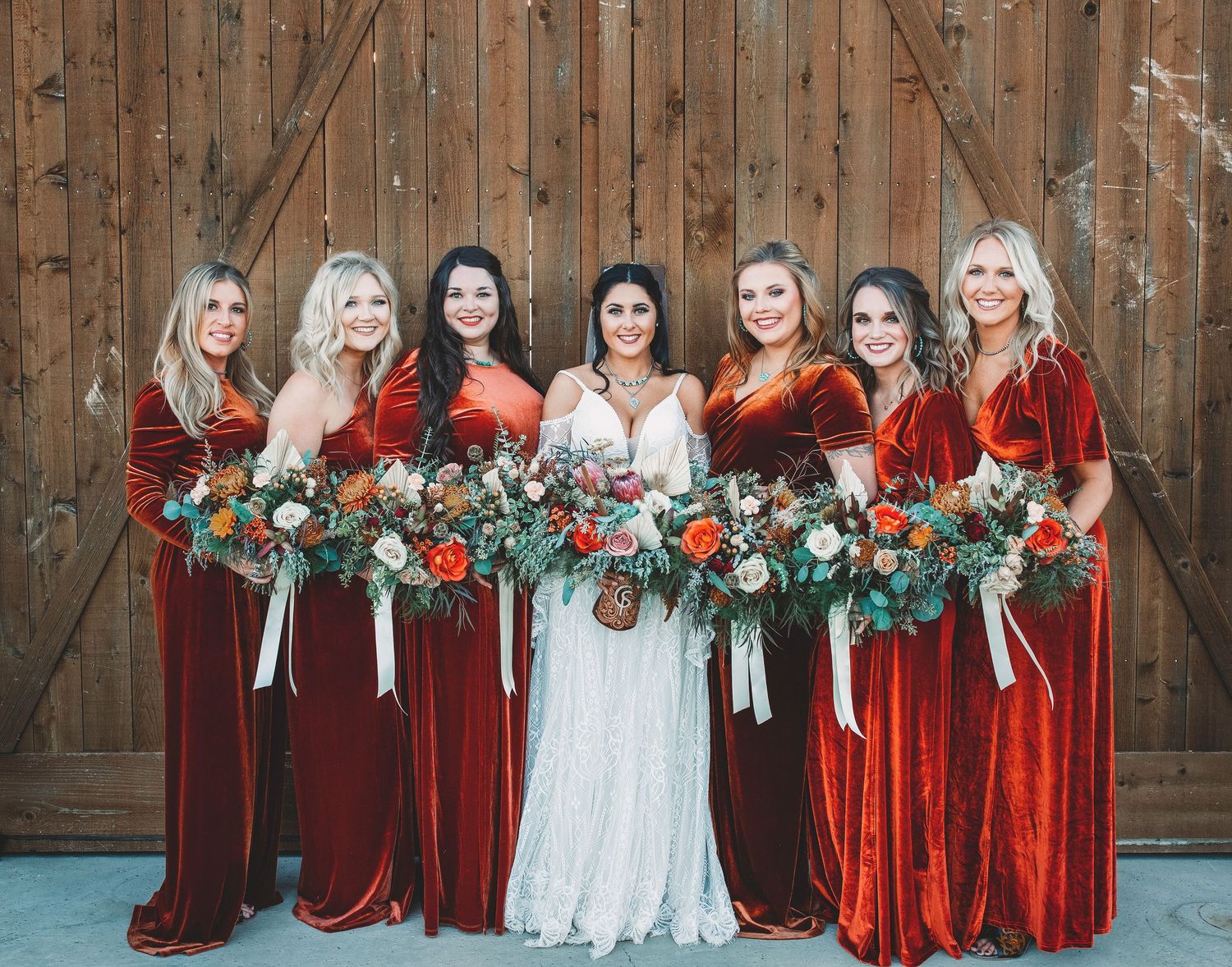 Wedding party in rich burgundy velvet dresses and a white bridal gown posing against a rustic wooden backdrop.