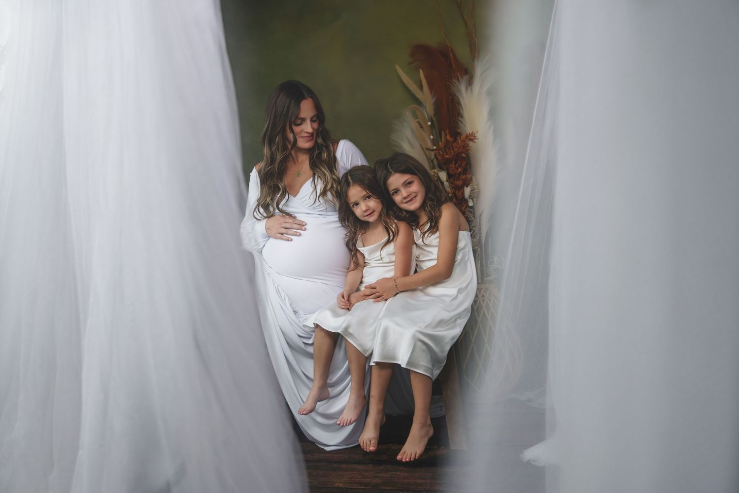 Three people in white dresses pose together between flowing white curtains in a dreamy portrait setting.