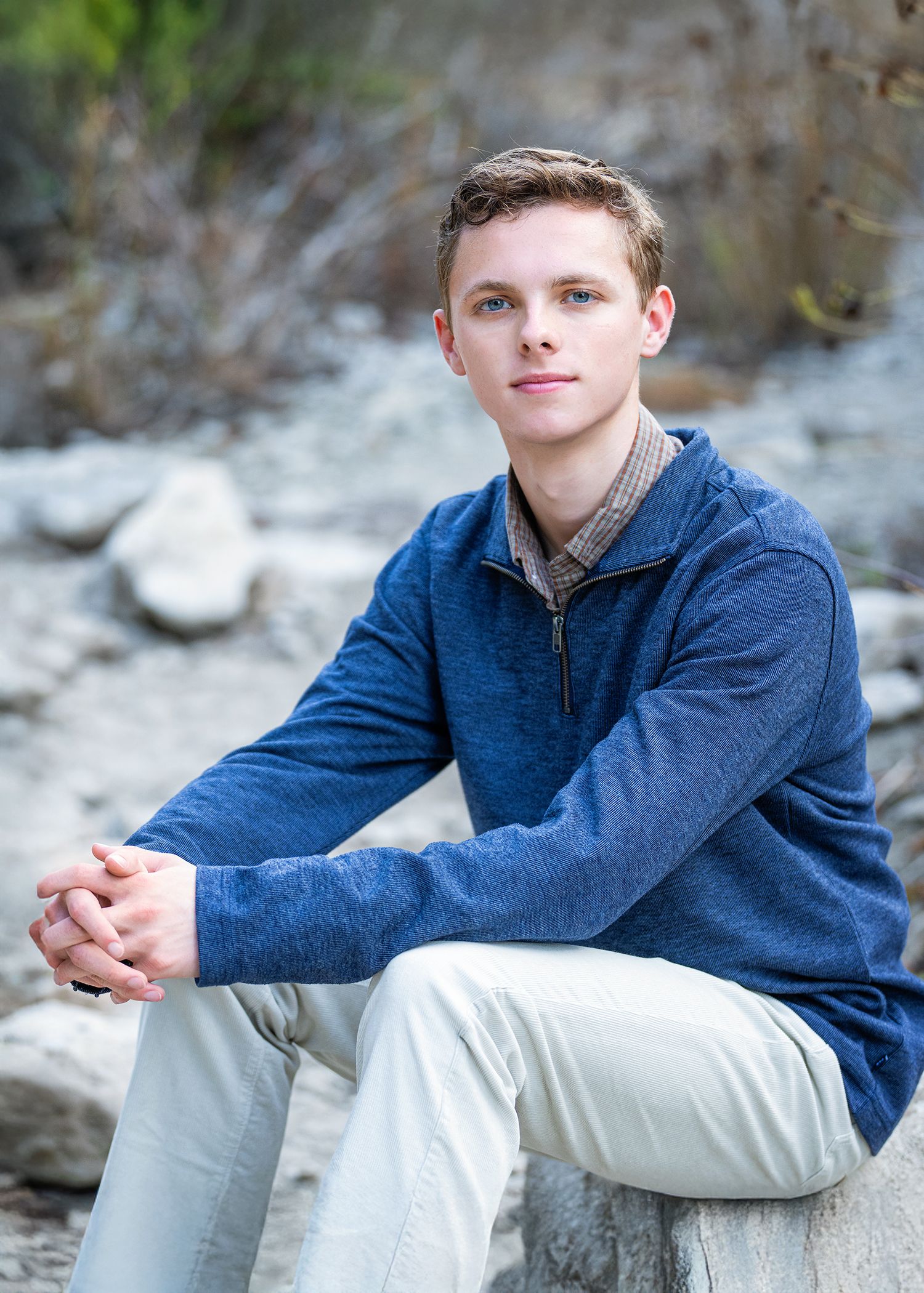 A person in a navy blue quarter-zip sweater and light pants sits casually on rocks in an outdoor setting.
