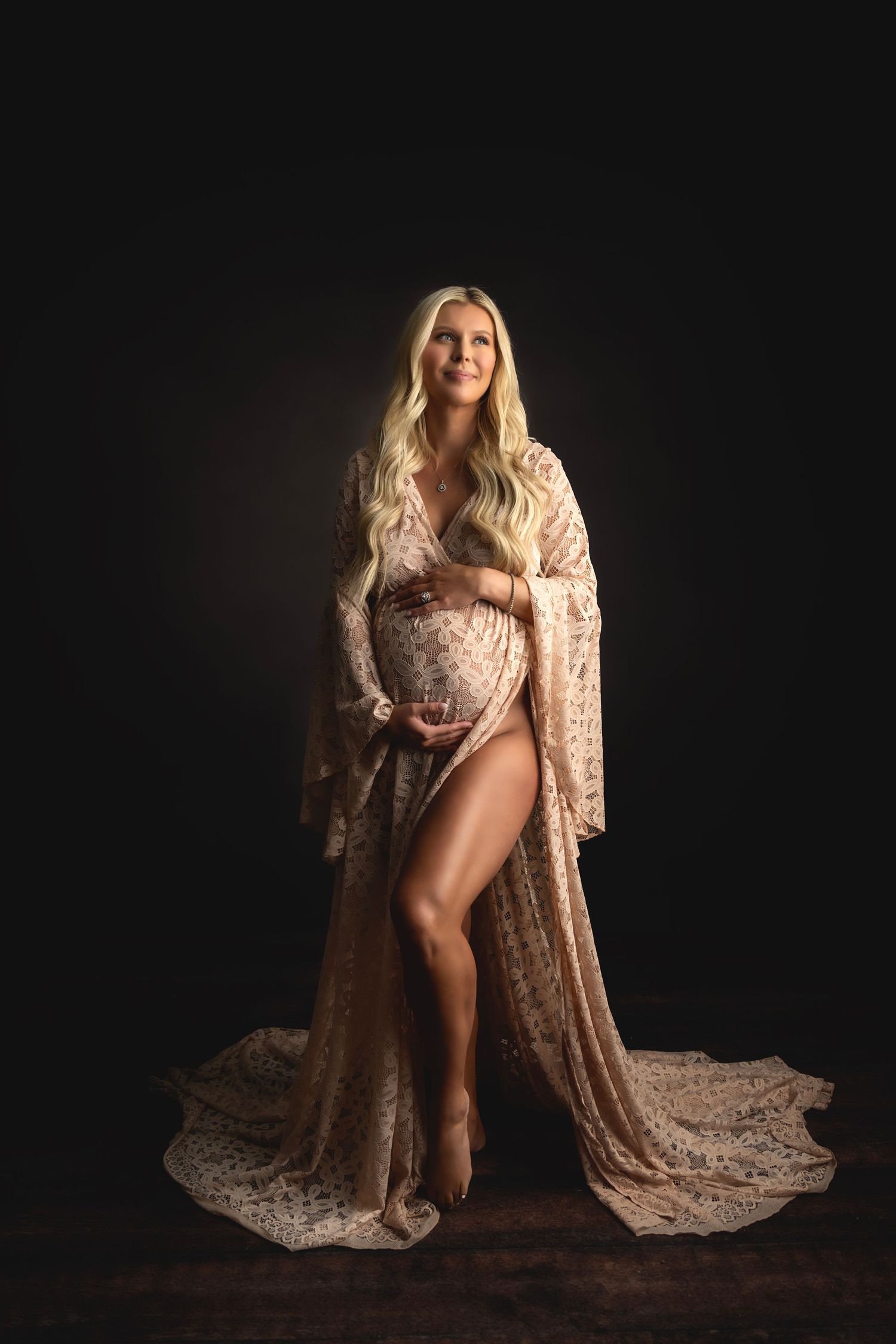 Glamorous maternity photo featuring woman in champagne lace gown against dark backdrop.