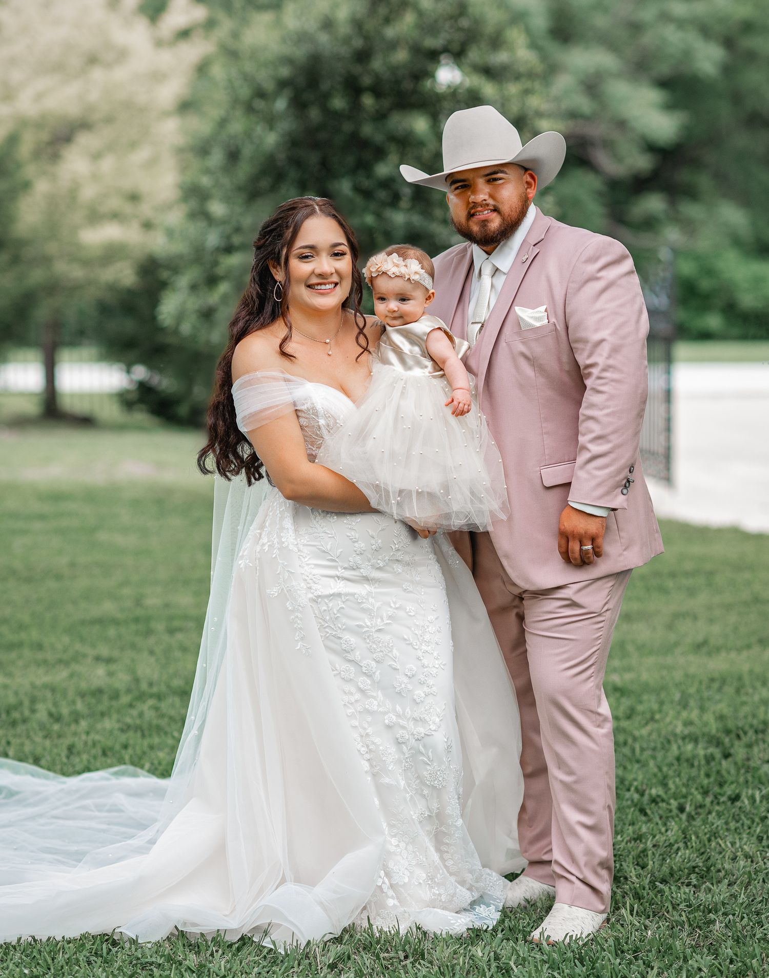 A romantic wedding photo on a grassy lawn featuring a couple in pink and white attire holding a small child.