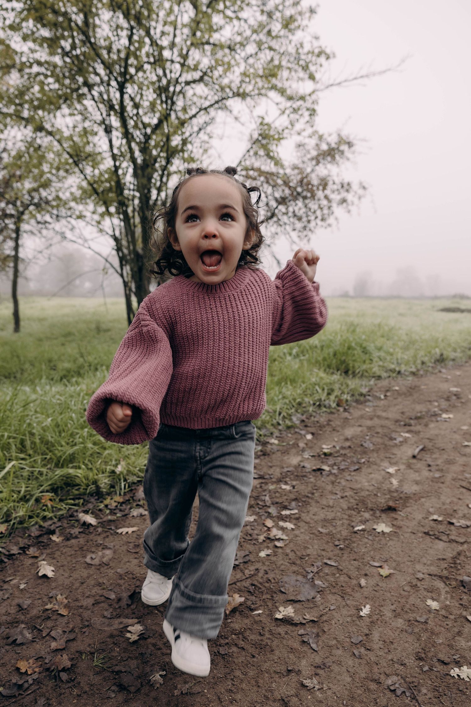 Small child in burgundy sweater and jeans runs joyfully along a dirt path on a misty autumn day.