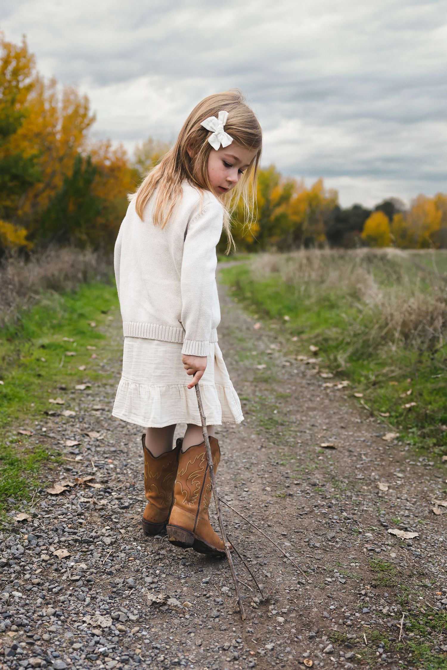 Child in white dress and brown boots walks along gravel path during fall season.