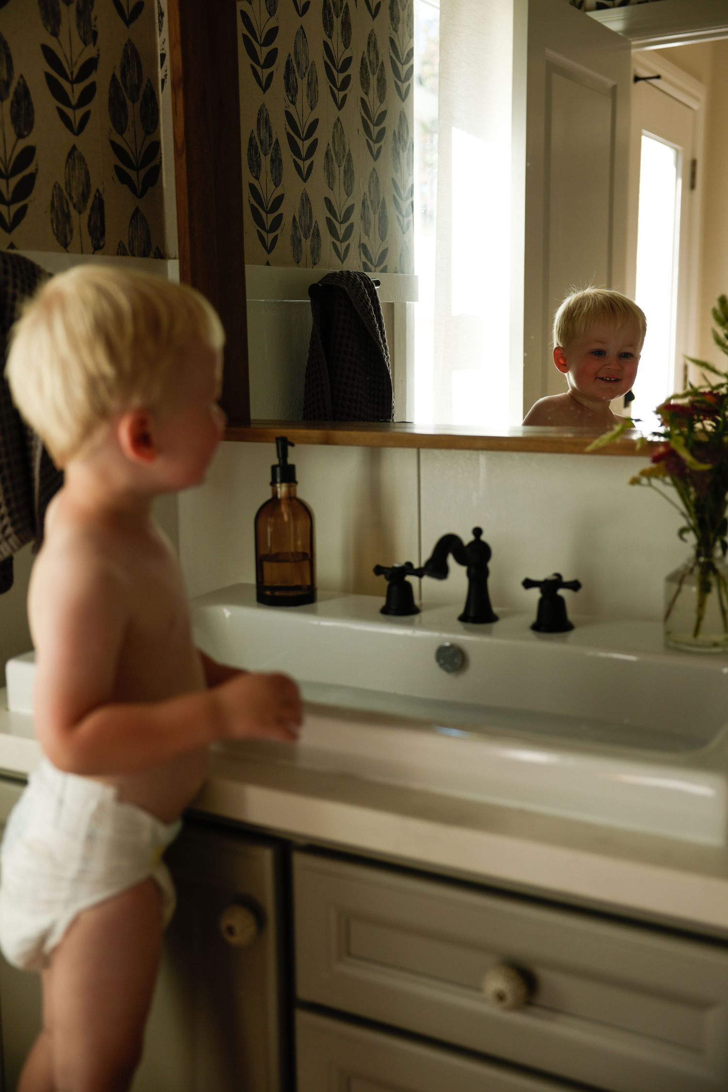 Looking in bathroom mirror with amber bottle and flowers on counter.
