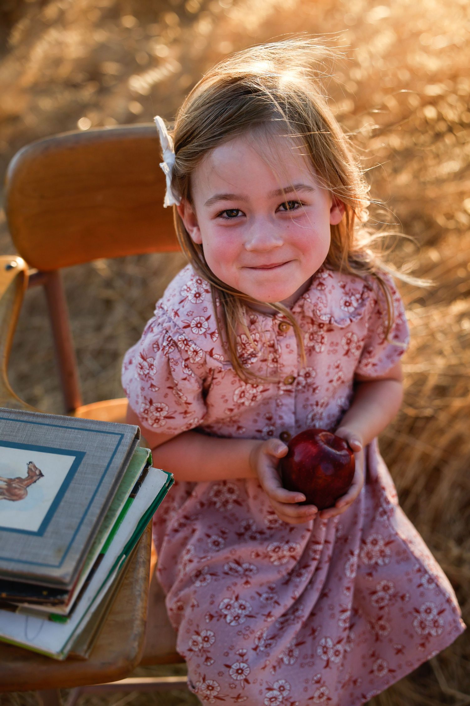 Child in floral dress explores books on rustic desk during golden hour photography session.
