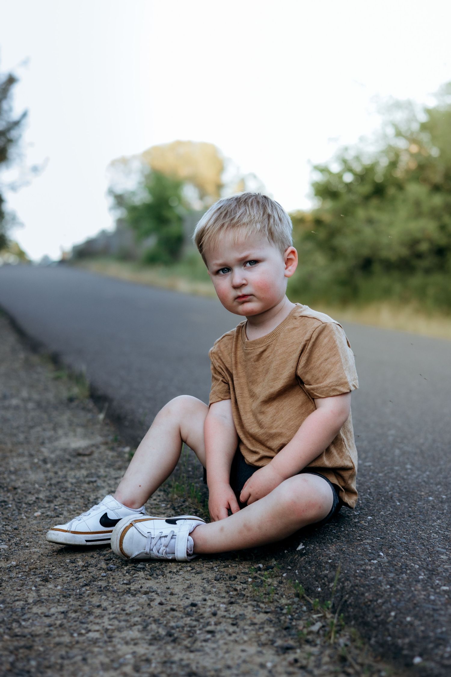 Young blonde boy in tan shirt and white Nike sneakers sitting on roadside edge with trees in background.