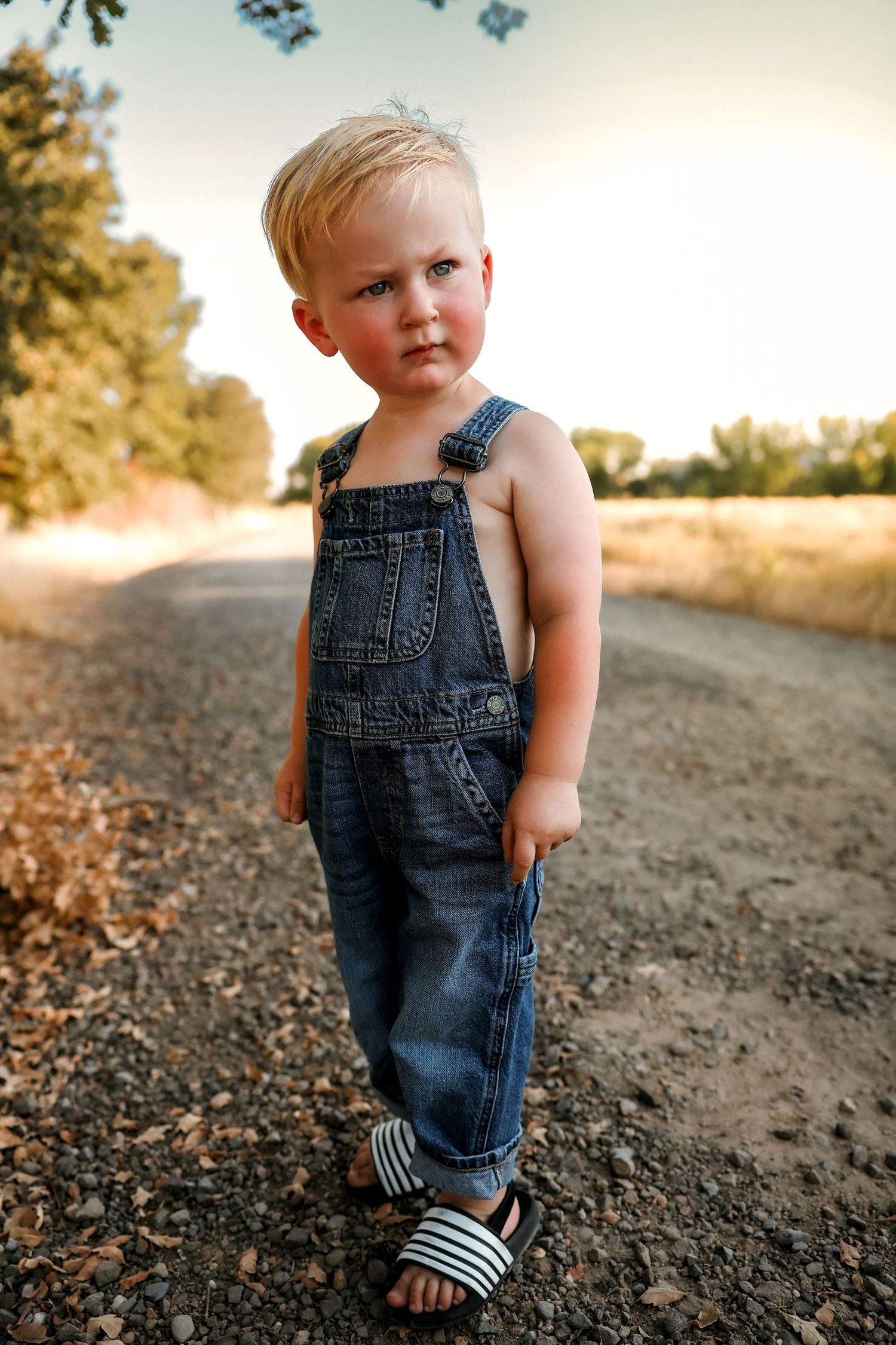 Small denim overalls and striped sandals stand on a dirt path at golden hour.