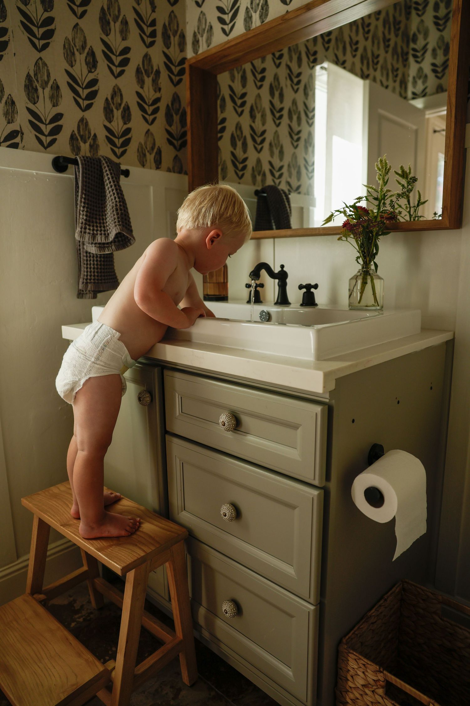 Standing on a wooden step stool at a gray bathroom vanity with patterned wallpaper.
