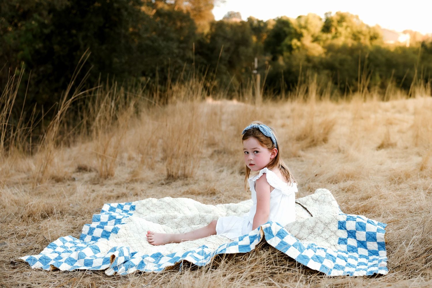 Children play on blue checkered blanket in dry grassy field during sunset.