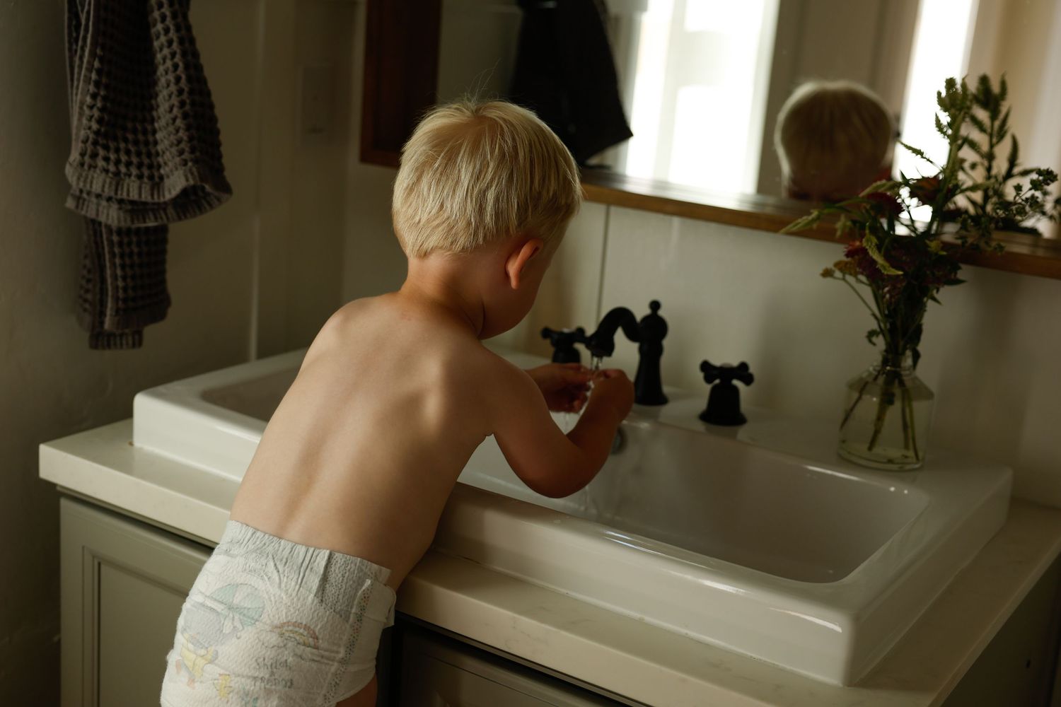 Playing at a bathroom sink with black faucet fixtures against patterned wallpaper.