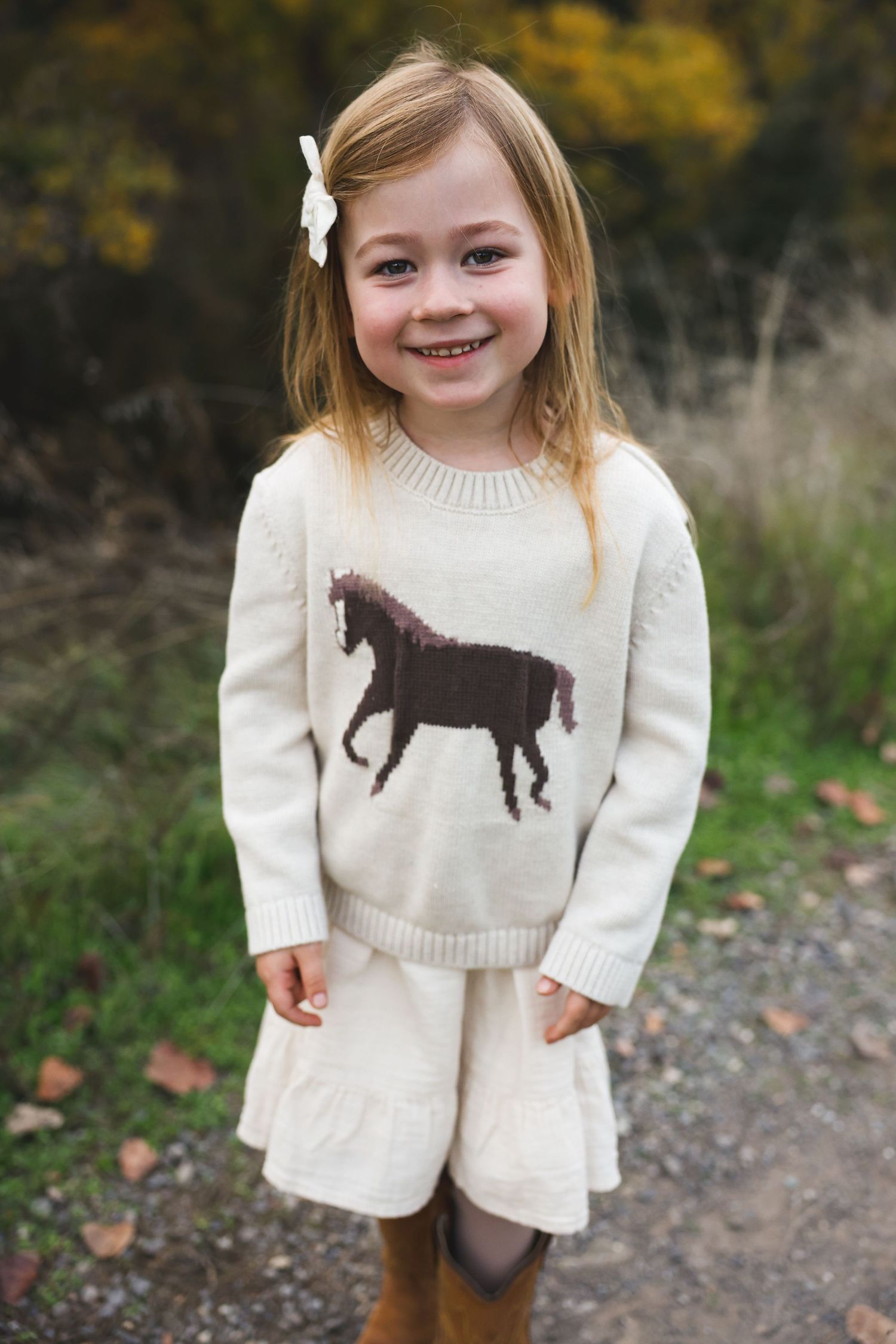Adorable white knit sweater with brown horse design shown in autumn outdoor setting.