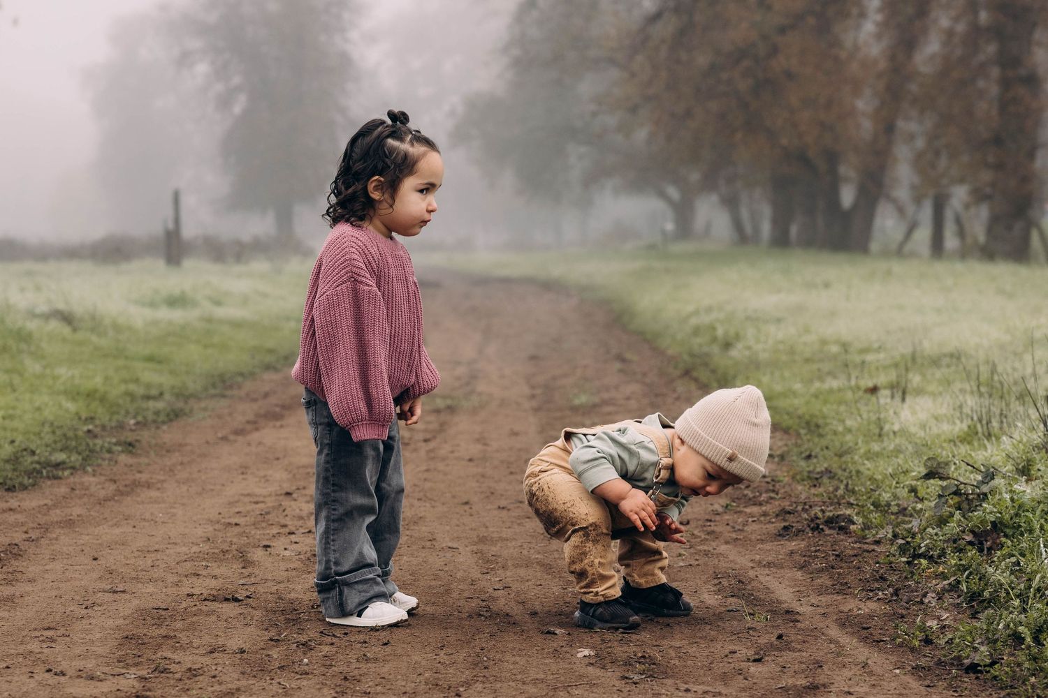 Children play together on a misty dirt path through a green field on a foggy morning.