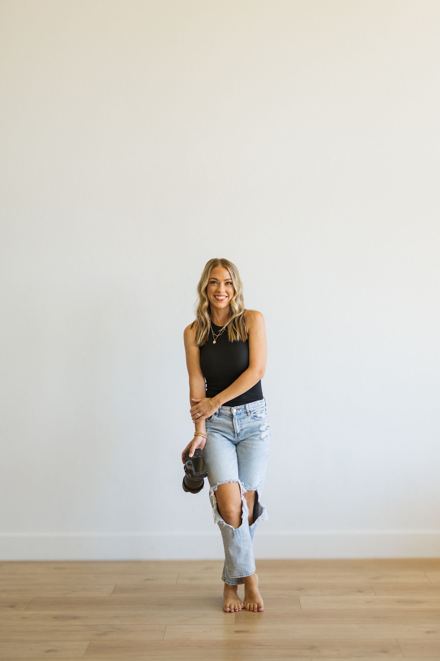 Smiling blonde woman in black tank top and ripped light wash jeans holding shoes, standing barefoot on hardwood floor.
