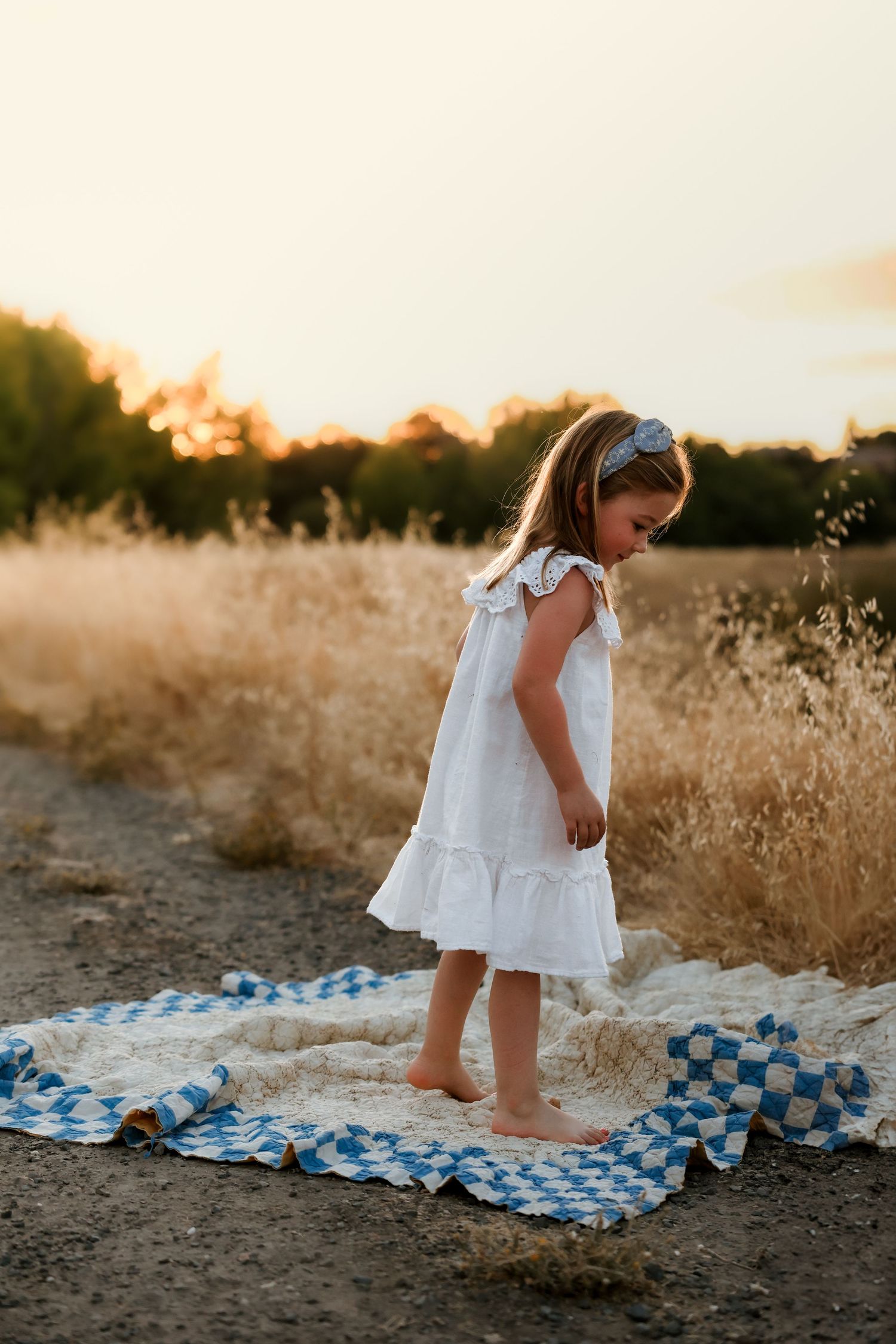A white dress-clad child standing barefoot on a blue checkered blanket at sunset.