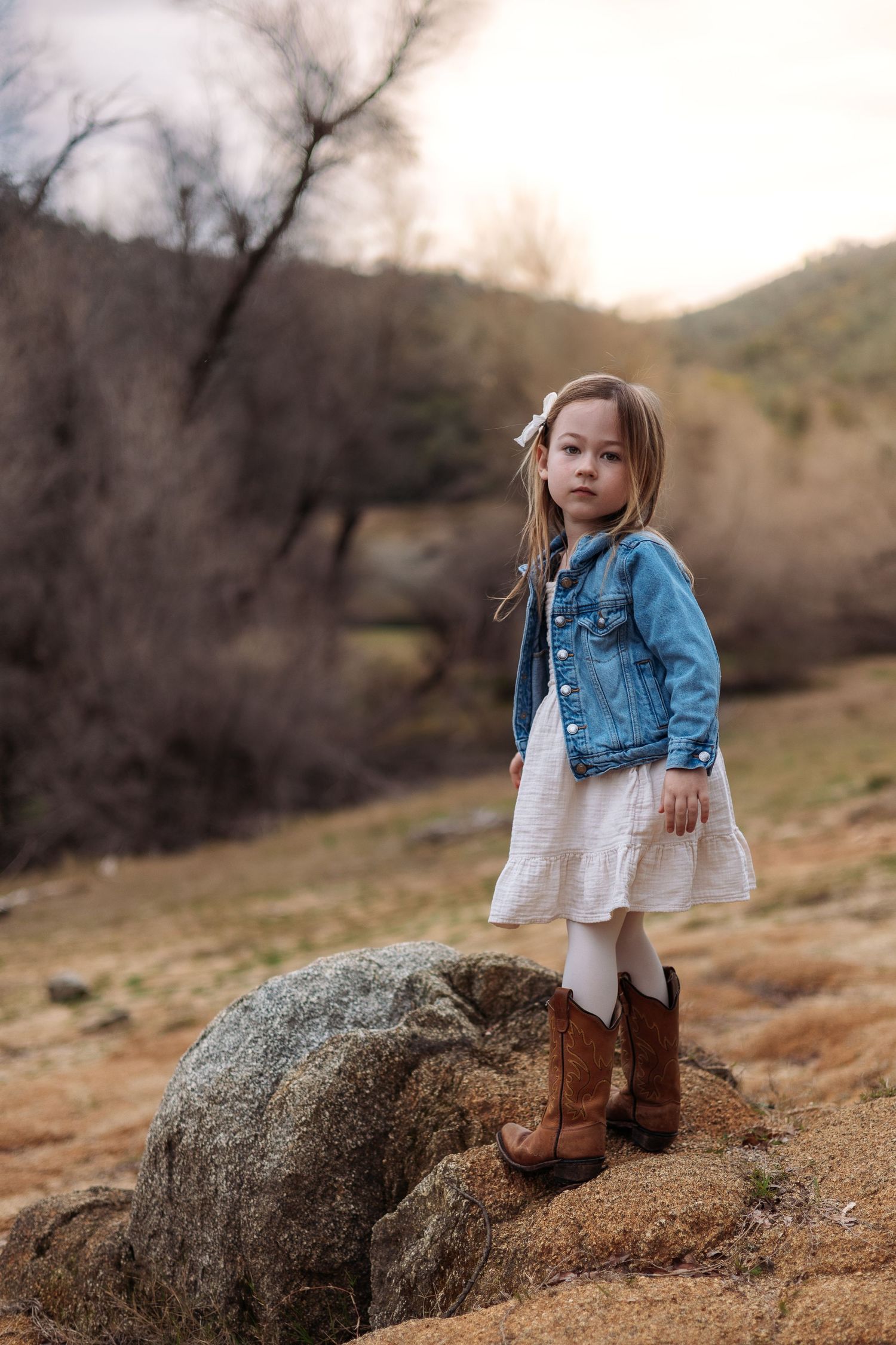 A young child in a denim jacket and white dress stands on a large rock in a natural outdoor setting during autumn.