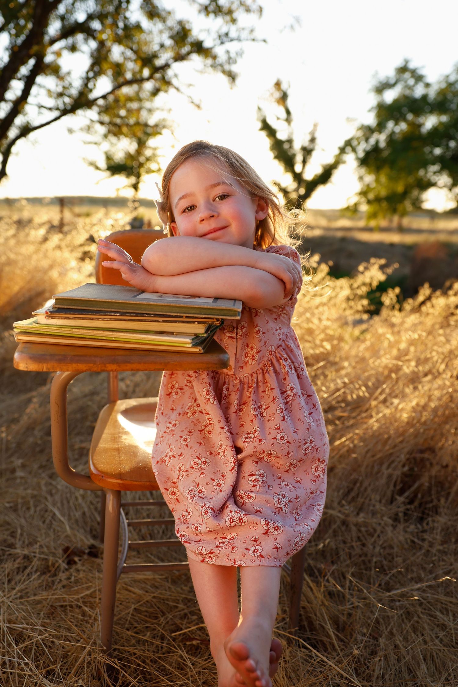 A small child in a pink polka dot dress sits at a wooden table with books during golden hour.