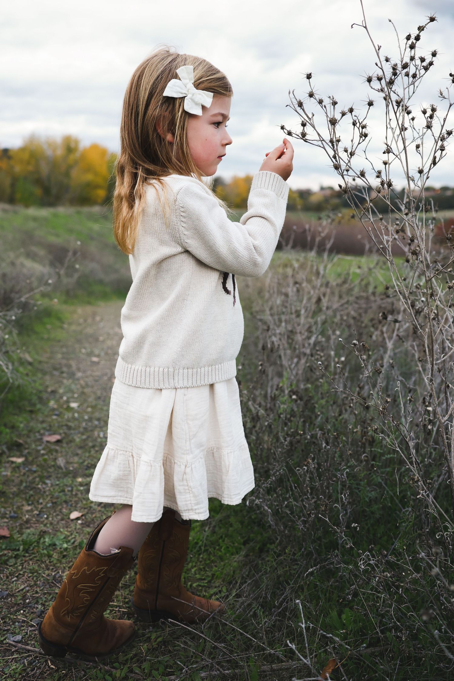 Child in white dress examines dried plants along a gravel path during fall.