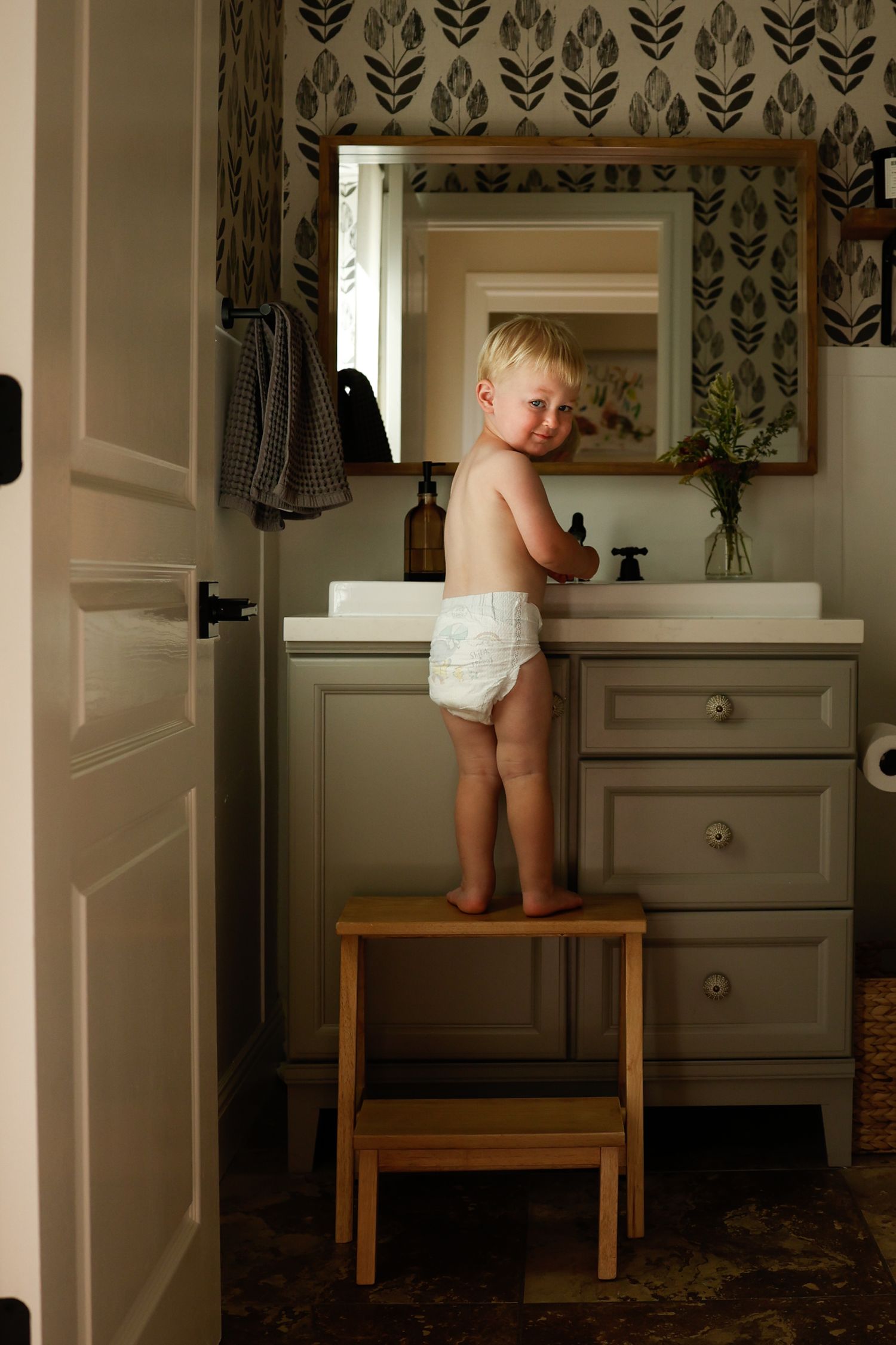 A bathroom scene showing a wooden step stool against a gray vanity with feather print wallpaper.