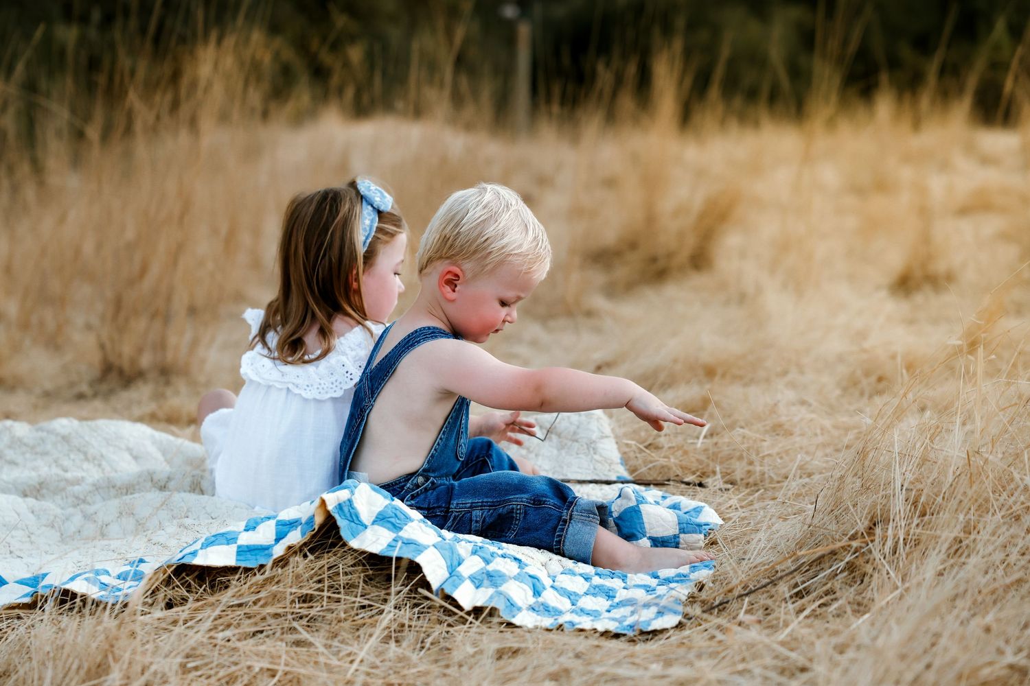 Children play together on checkered blanket in grassy field at sunset.