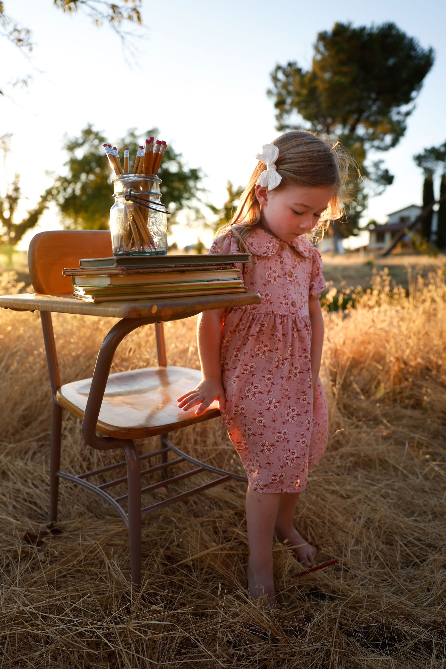 A pink floral dress and antique wooden chair create a whimsical scene in golden sunlit grass at sunset.