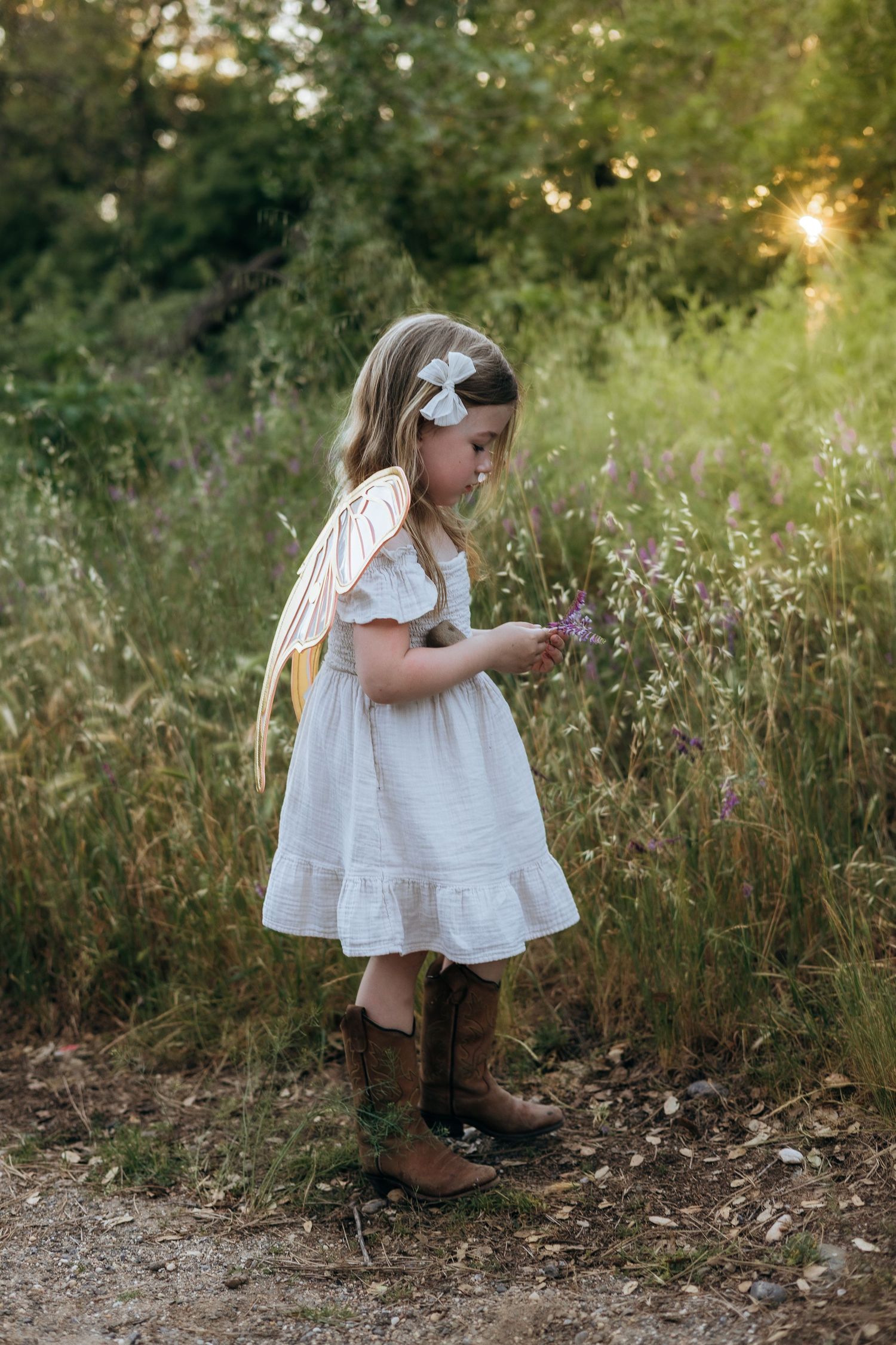 Young girl in white dress and cowboy boots picks wildflowers at golden hour, wearing fairy wings and bow headband.