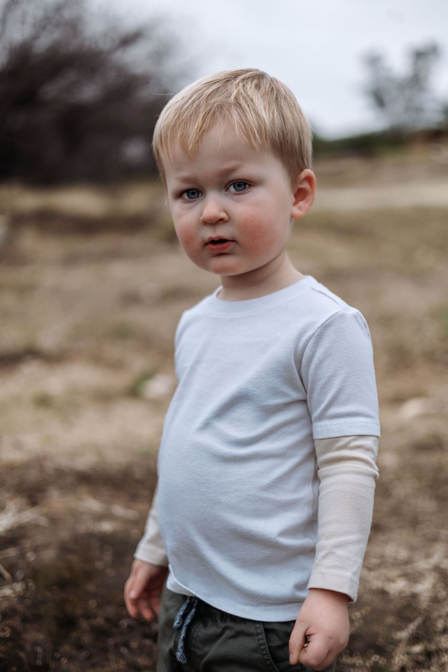 A toddler in a white t-shirt stands outdoors against a blurred natural background.