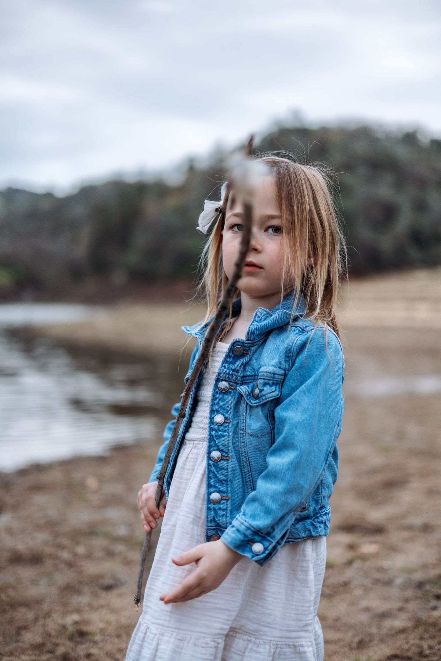 A young child in a denim jacket and white dress stands on a beach looking up at the sky with wonder.