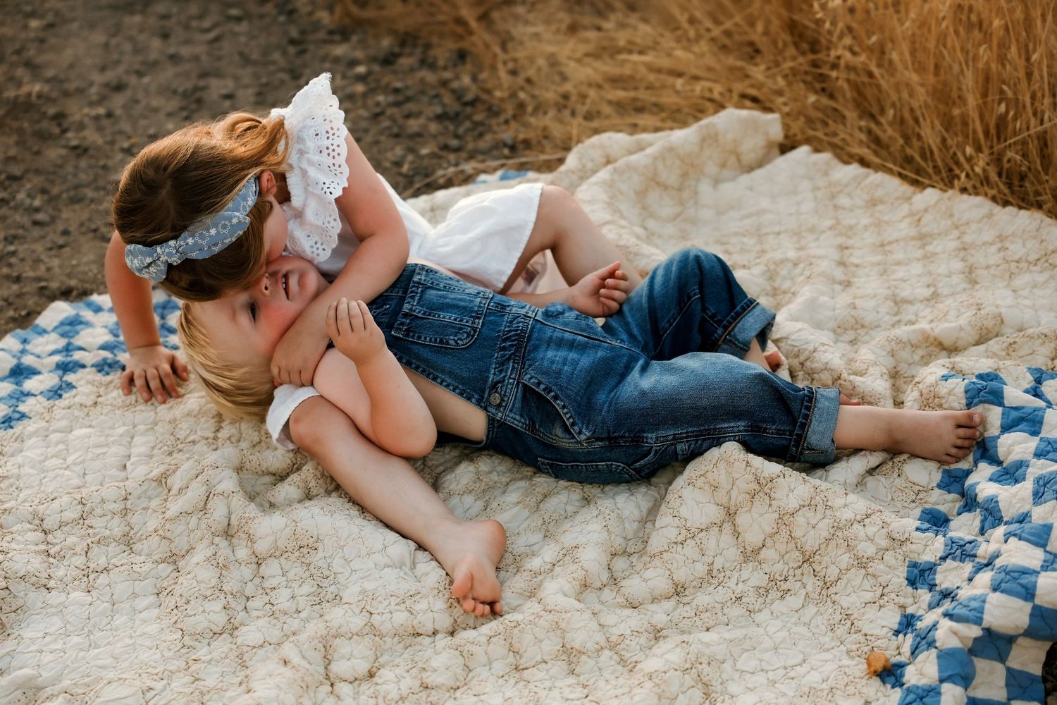 Two children playfully resting together on a patterned blanket outdoors during sunset.