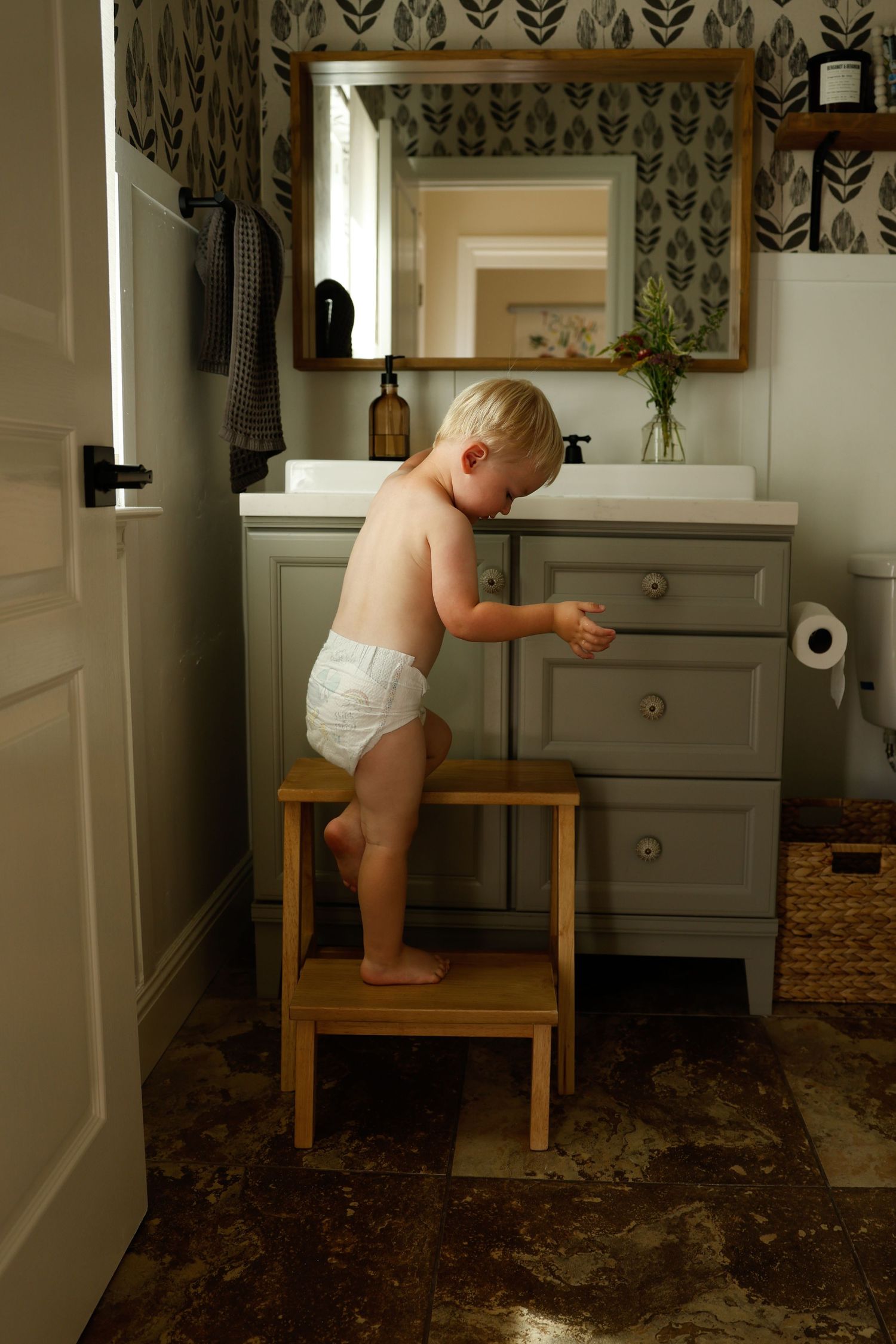 A toddler in a diaper climbs on wooden furniture in a bathroom with patterned wallpaper.
