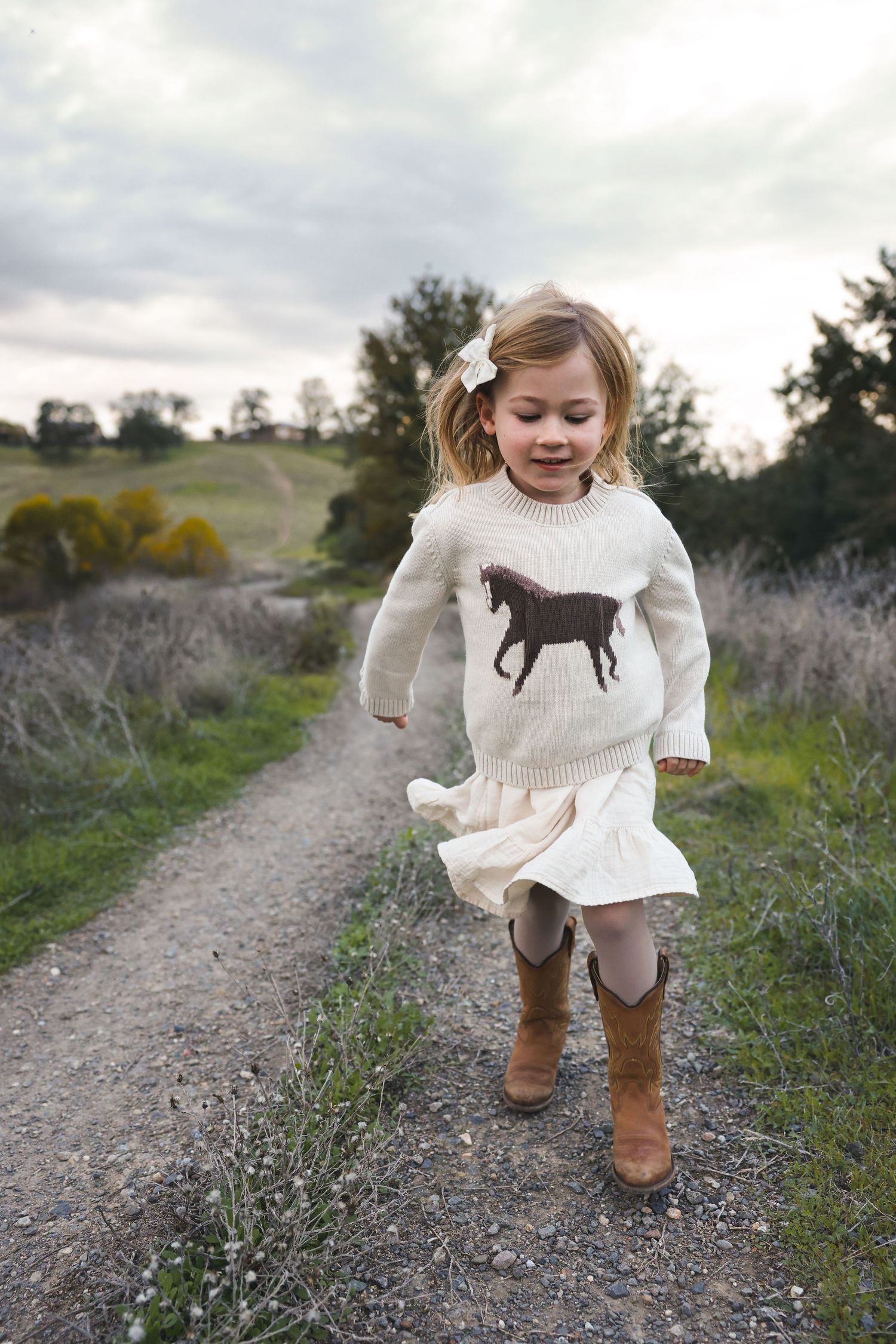 Child in white sweater with horse design jumps playfully on a countryside path.