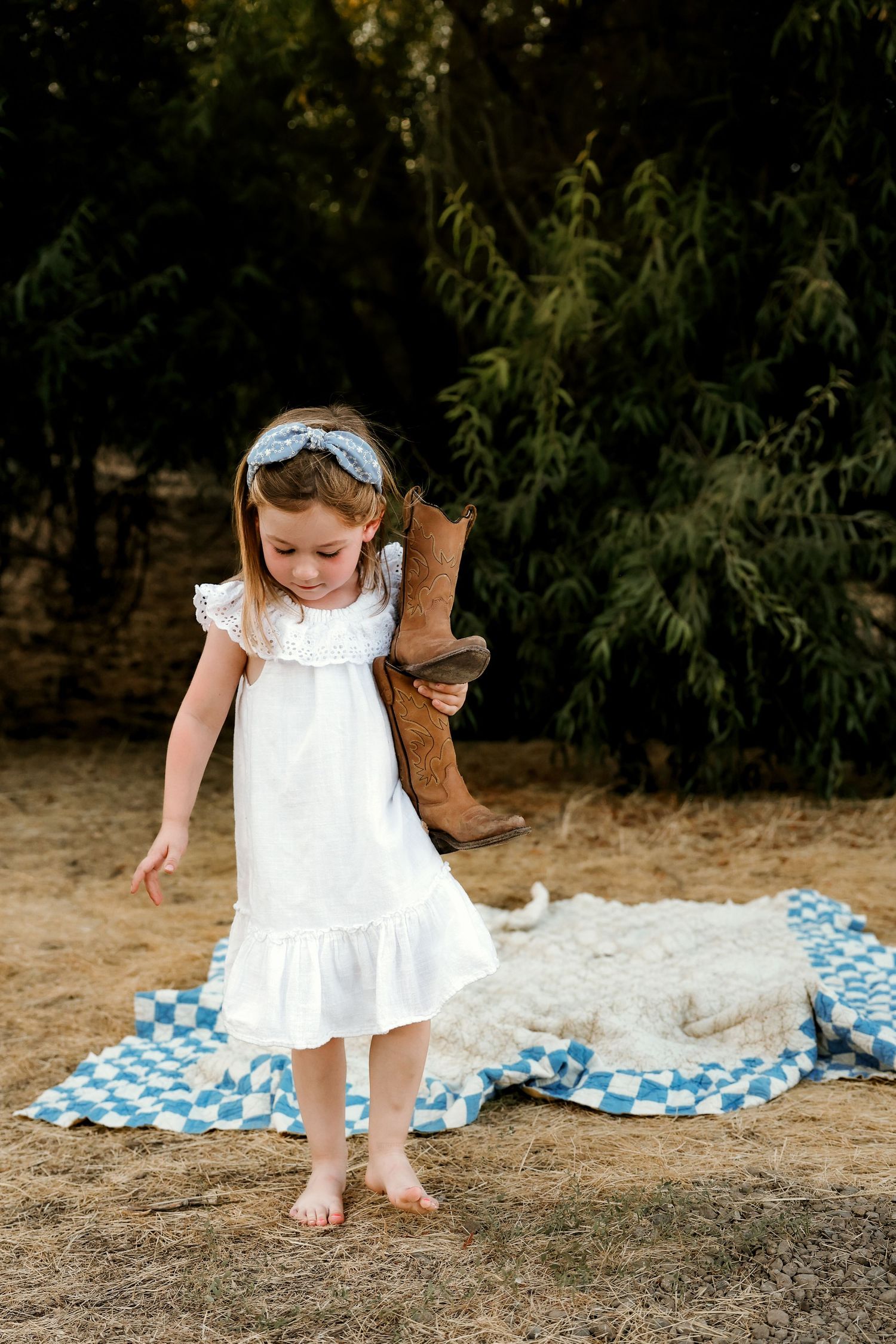 Young child in white dress explores outdoor setting with blue checkered blanket nearby.