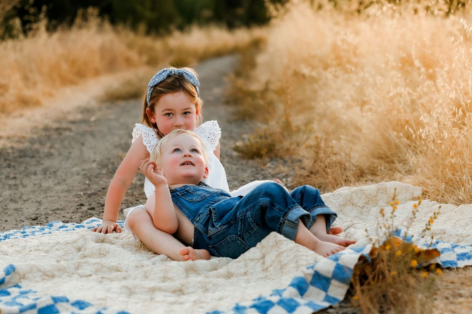 Two children playing together on a blue and white checkered blanket during golden hour on a dirt path.