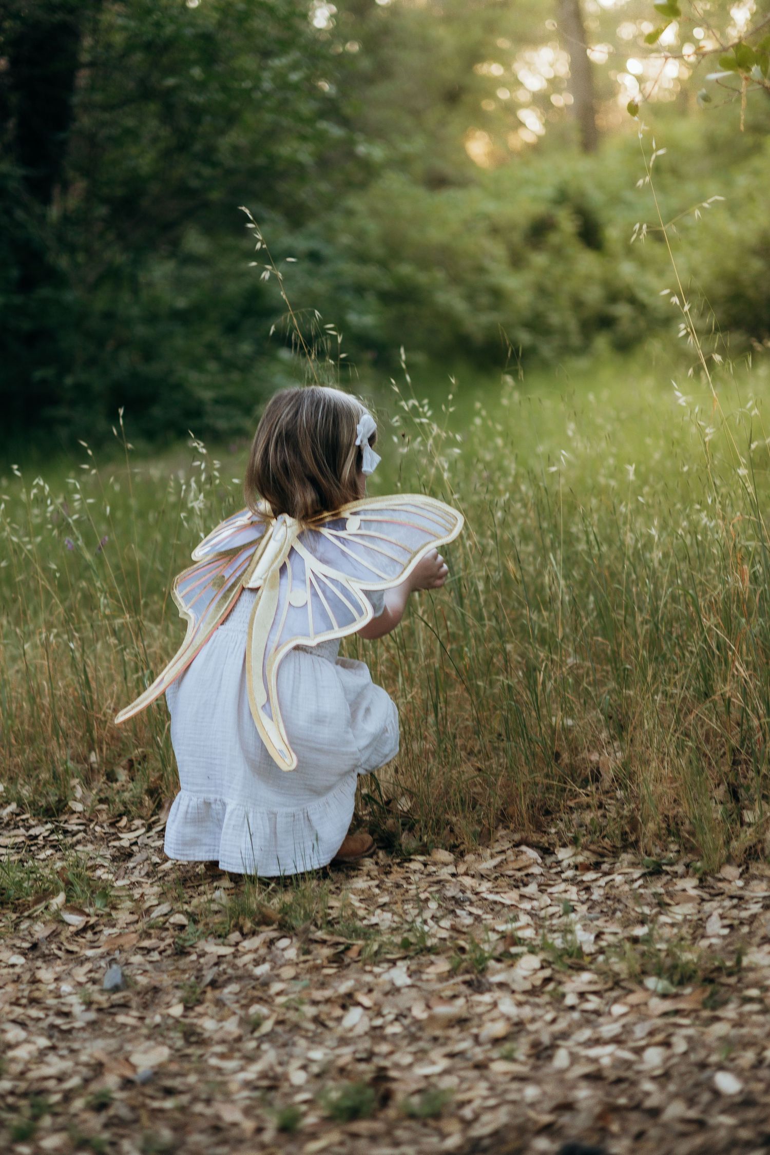 Young girl in white dress with fairy wings crouching in a forest meadow during golden hour.