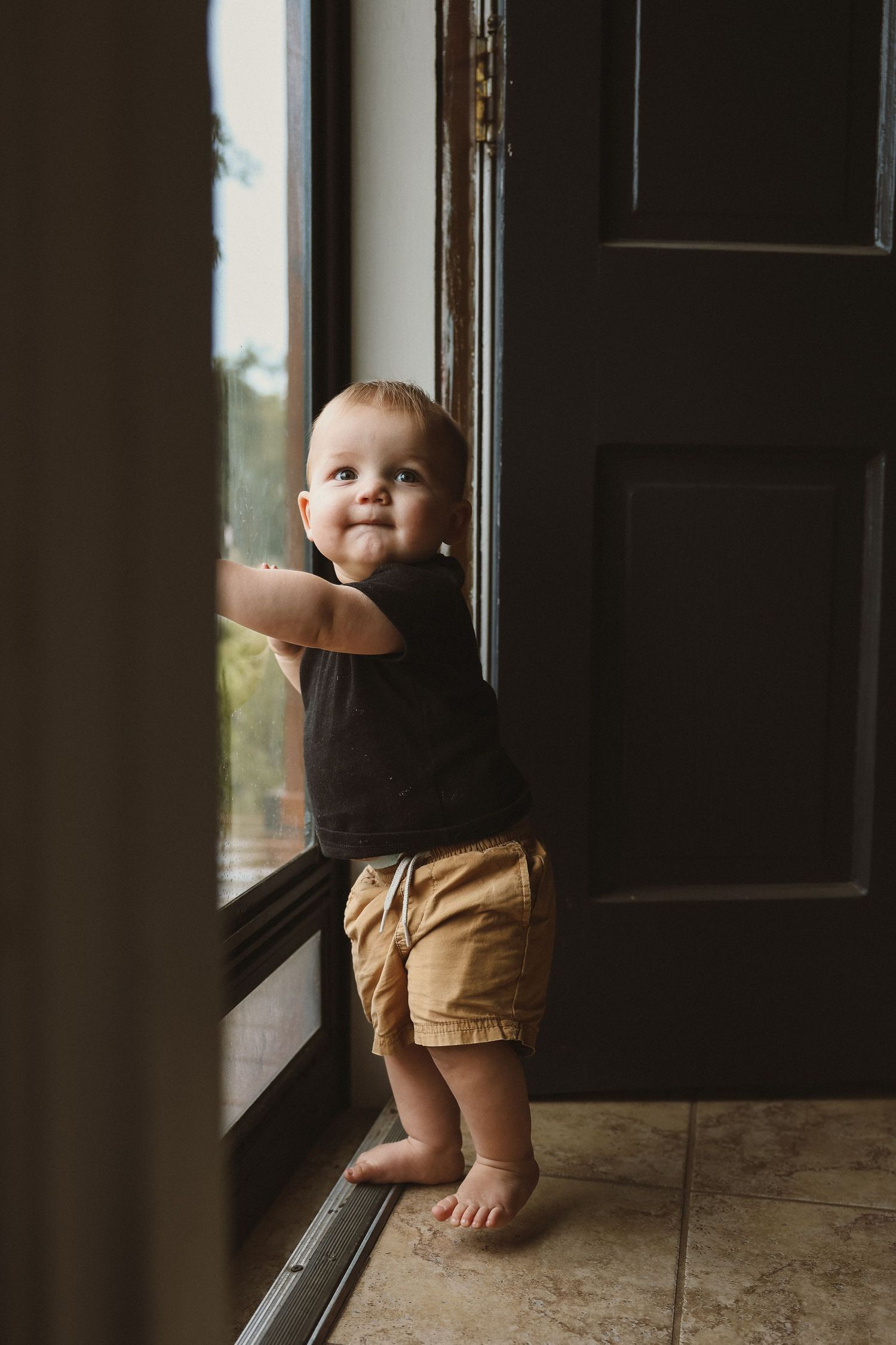 A baby in brown shorts peers through a glass door in moody indoor lighting.
