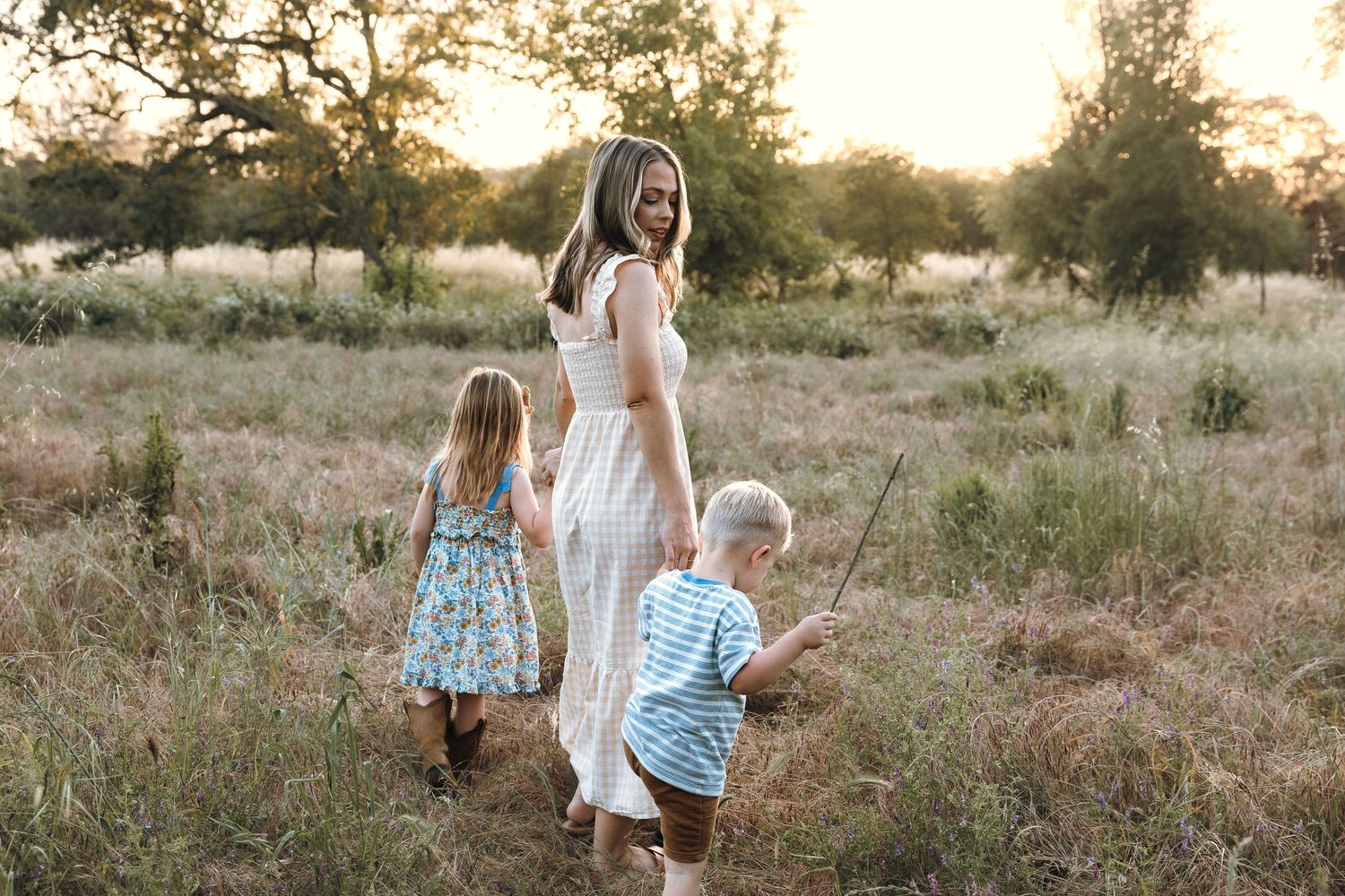 A mother in a white dress walks through a golden field with her two young children at sunset.