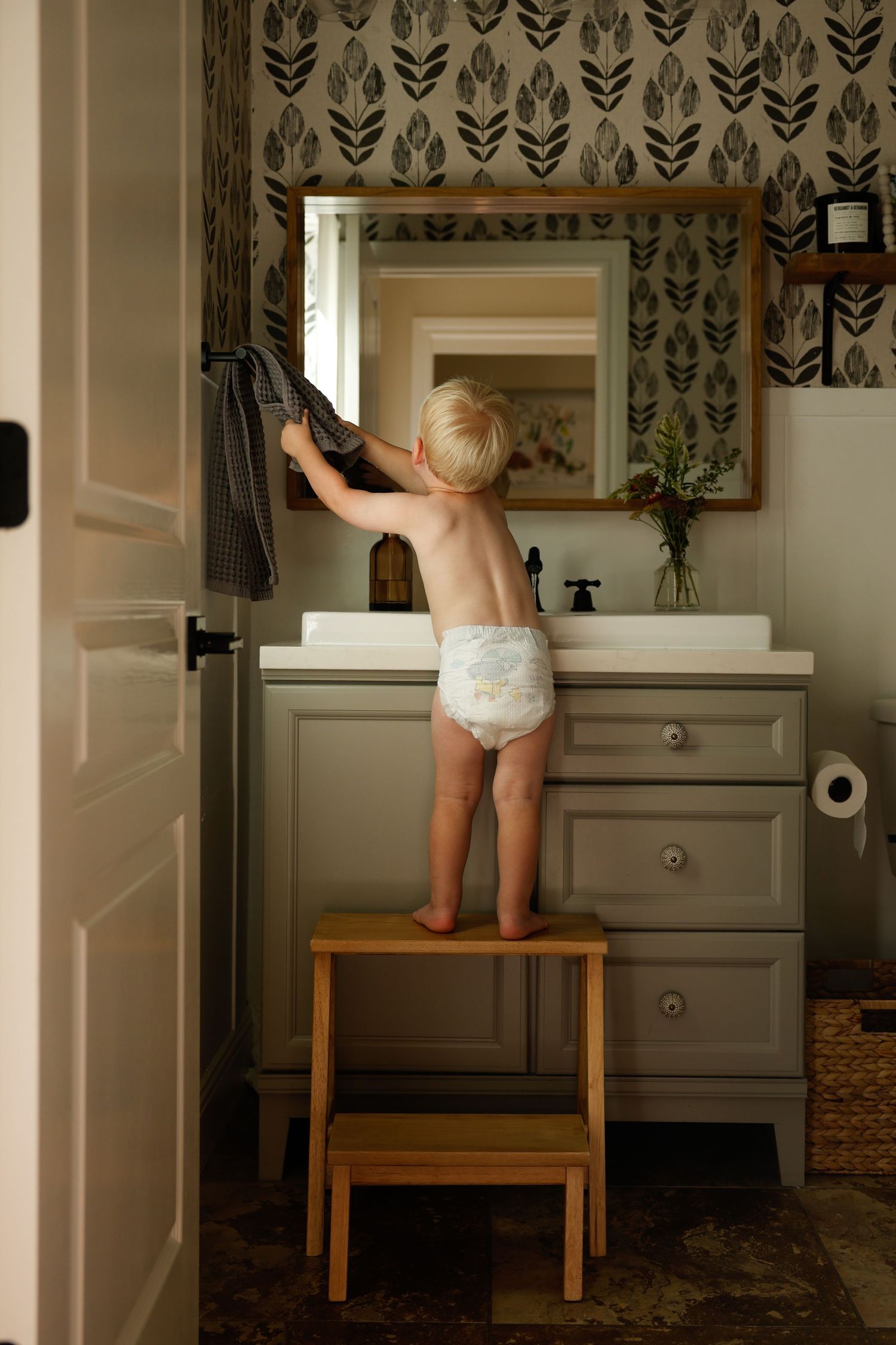 Standing on a wooden step stool reaching up to the bathroom mirror.