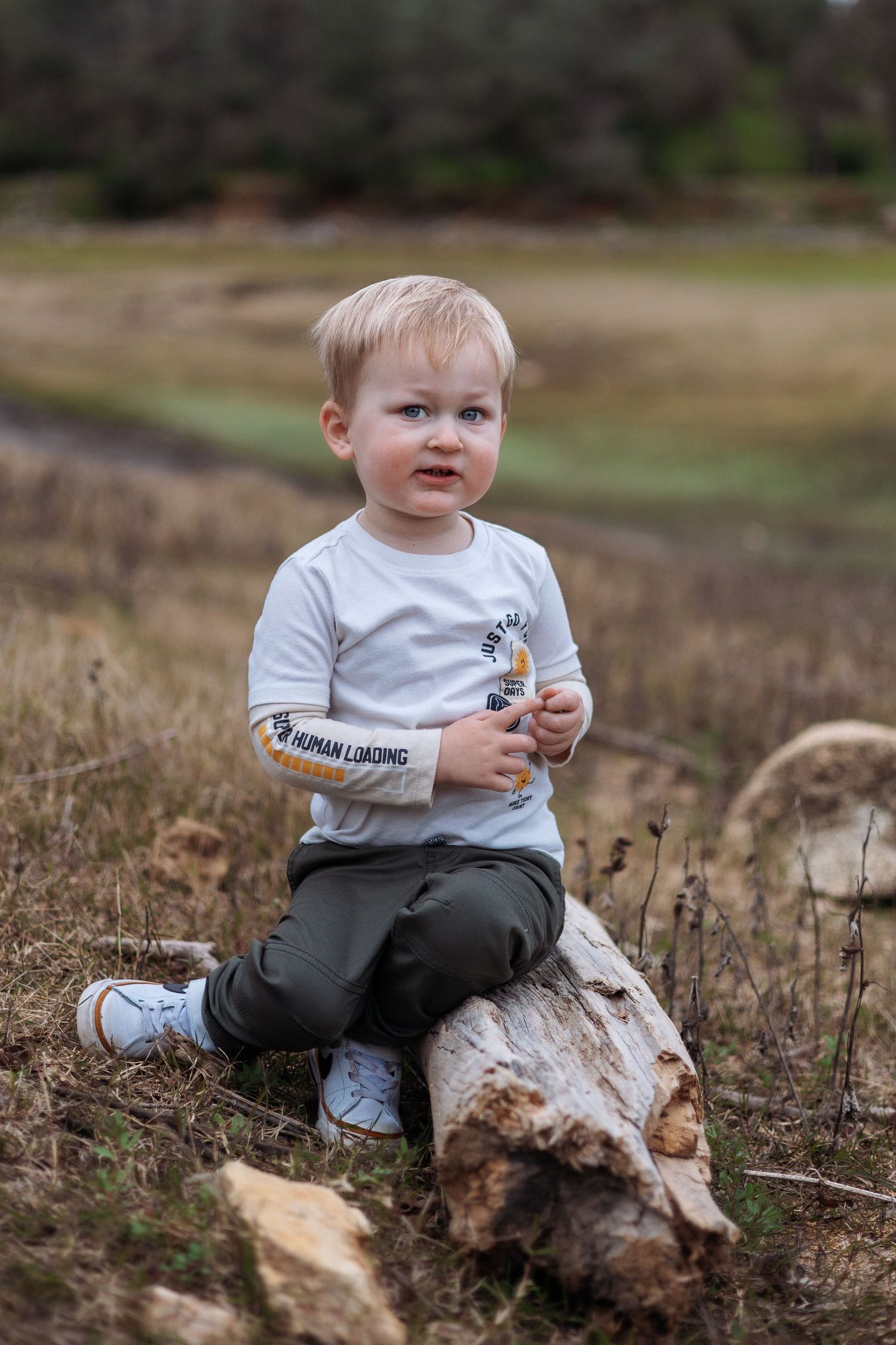 A toddler in a white shirt and gray pants sits on a rustic log in an outdoor field setting.