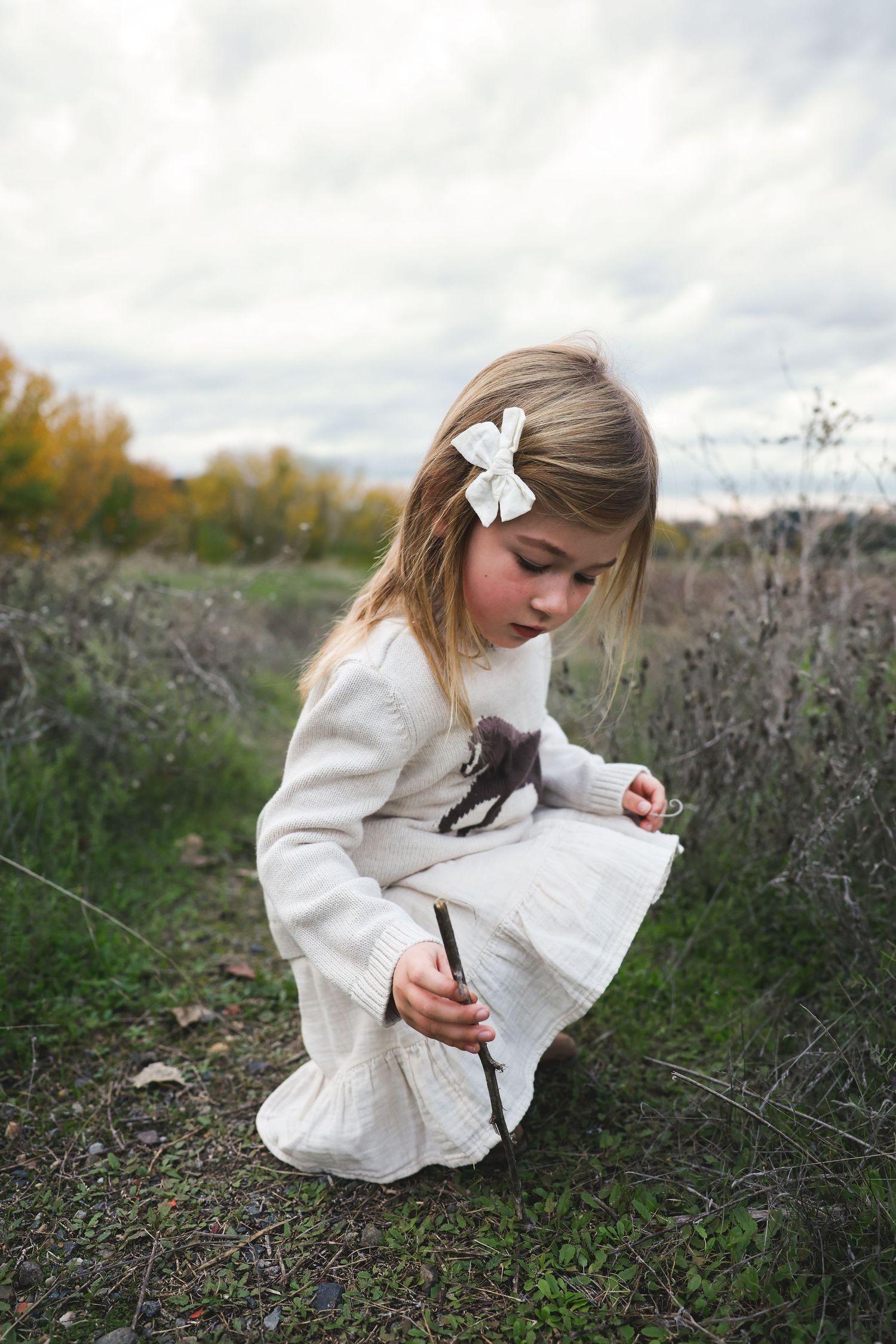 Child in cream dress crouches to examine dried plants along a nature path.