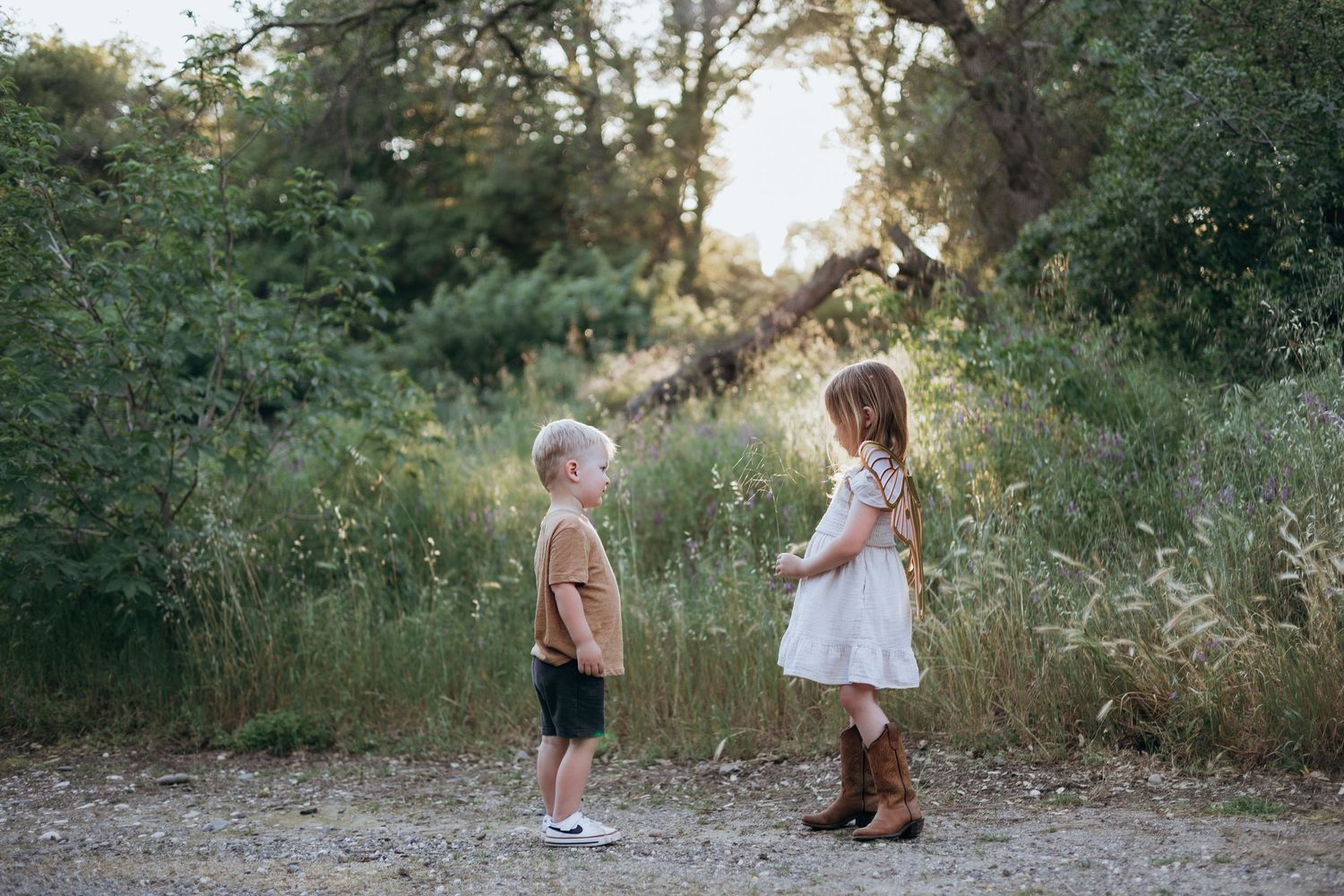 Two young children face each other on a dirt path surrounded by lush green trees in a warm outdoor setting.