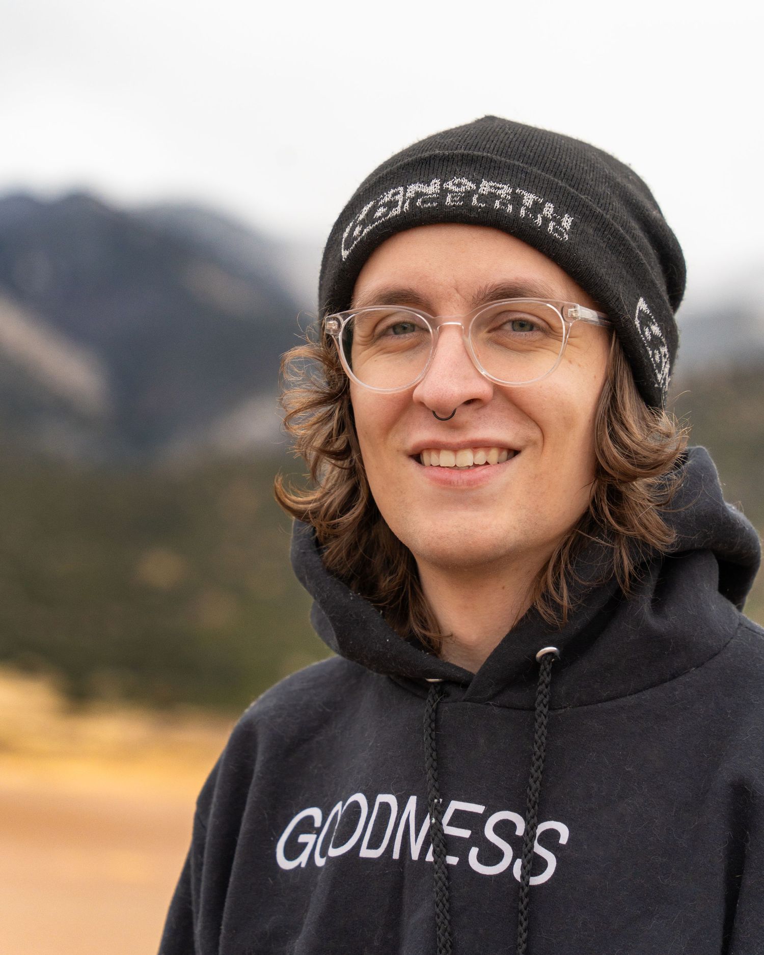 Person wearing a black hoodie with 'Goodness' text smiles outdoors against a mountain backdrop.