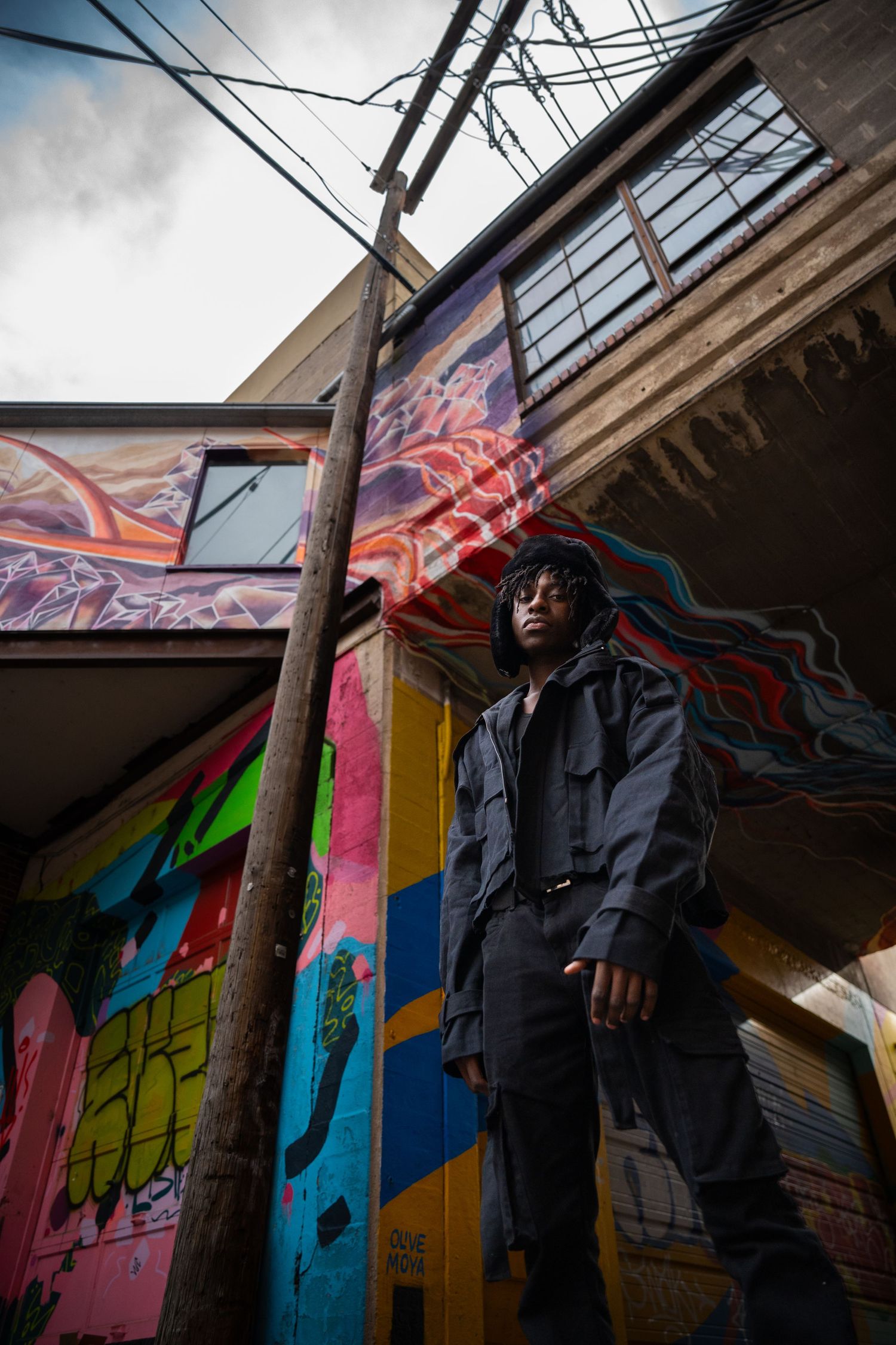 Portrait in front of urban street art mural with vibrant colors. Backdrop includes near power lines and building exterior.
