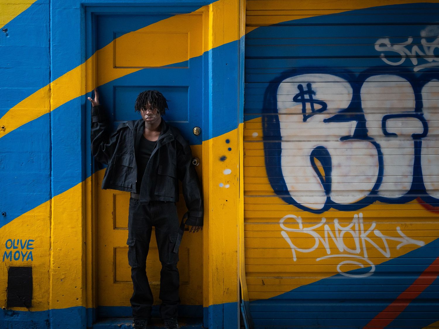 Portrait in front of a yellow and blue striped wall with graffiti art and shadows creating an urban geometric pattern.