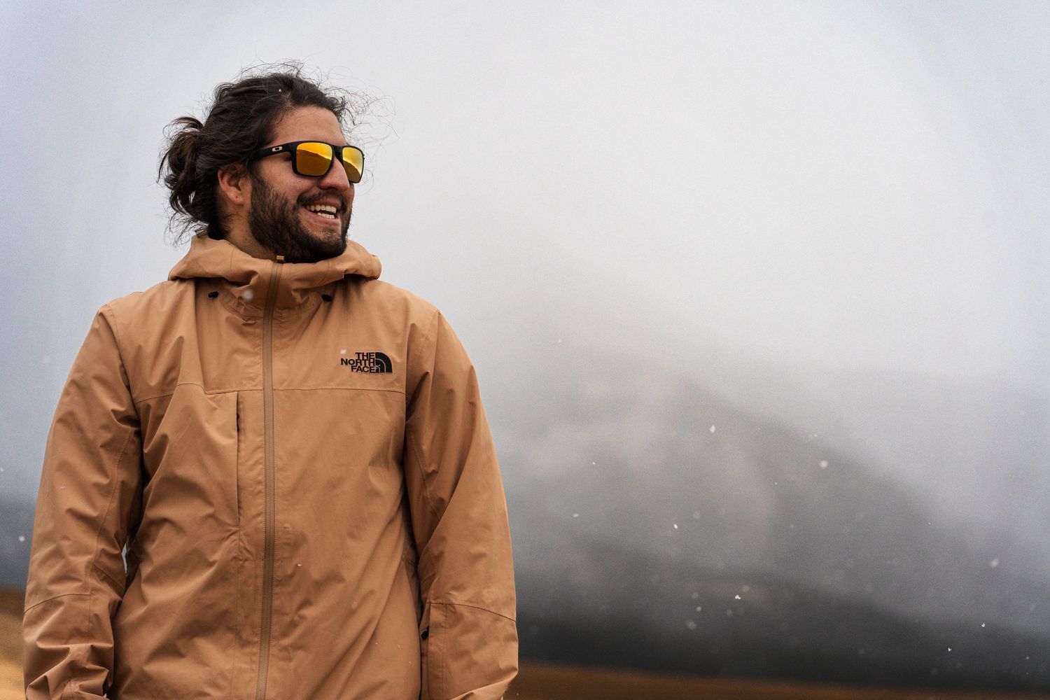 Person wearing tan North Face jacket smiles against misty mountain backdrop.