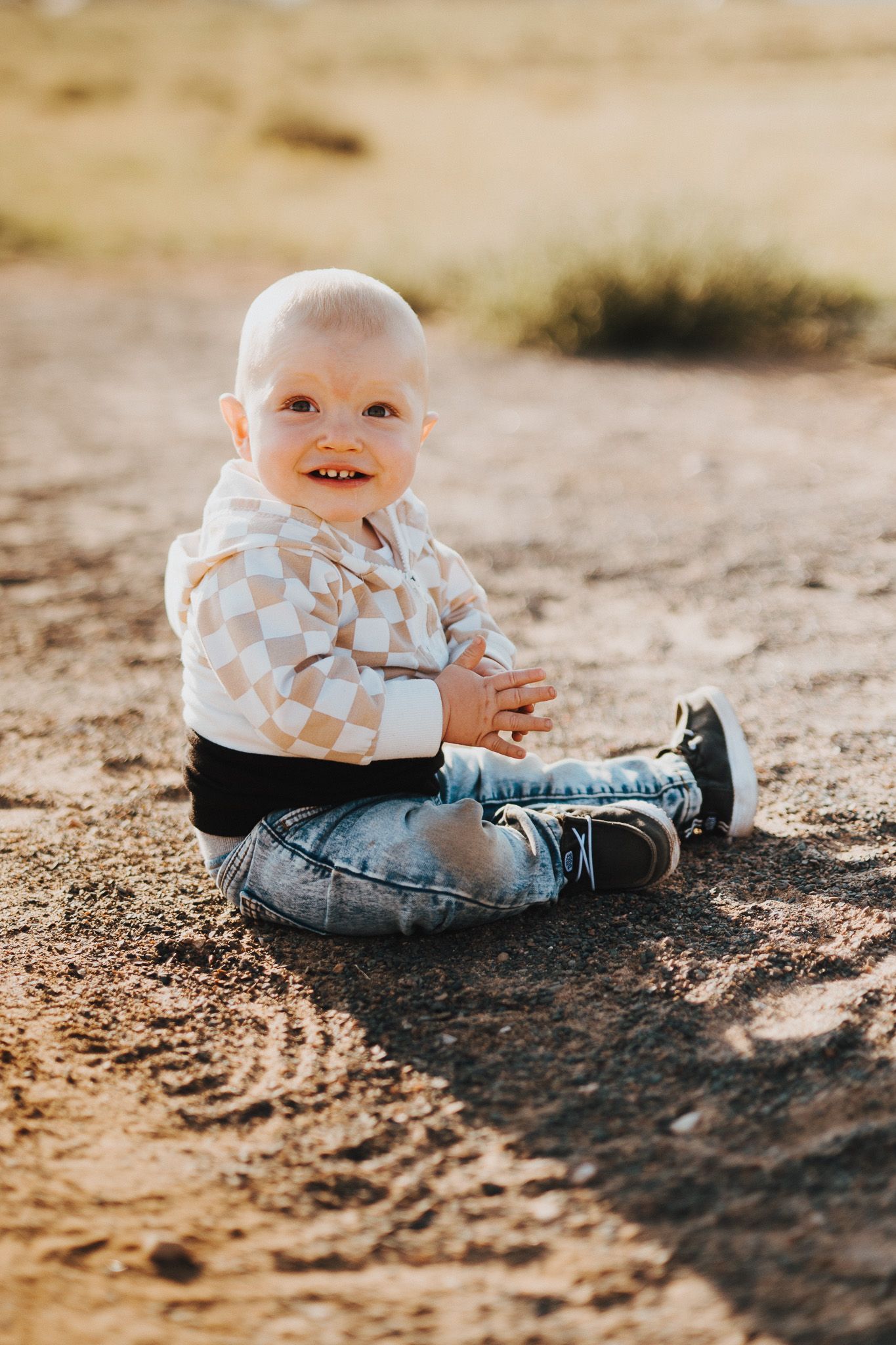 Small child in blue and white patterned shirt sits and plays in sandy dirt on sunny day.