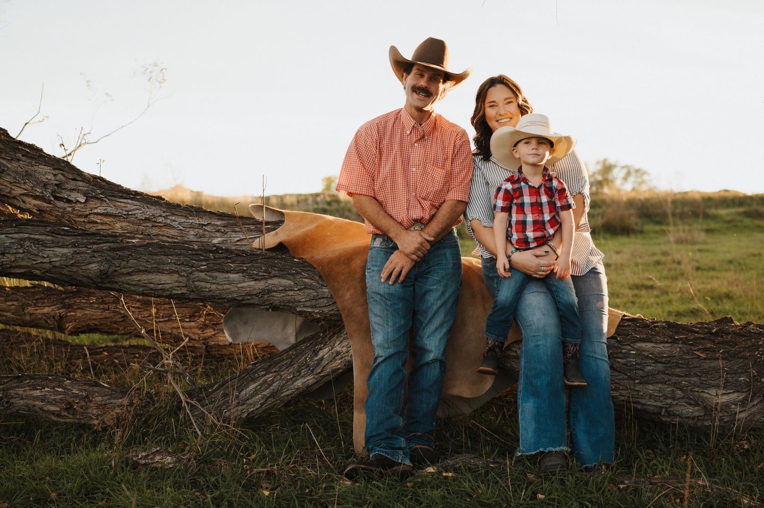 A couple in western attire sits together on a fallen log at sunset in a rustic outdoor setting.