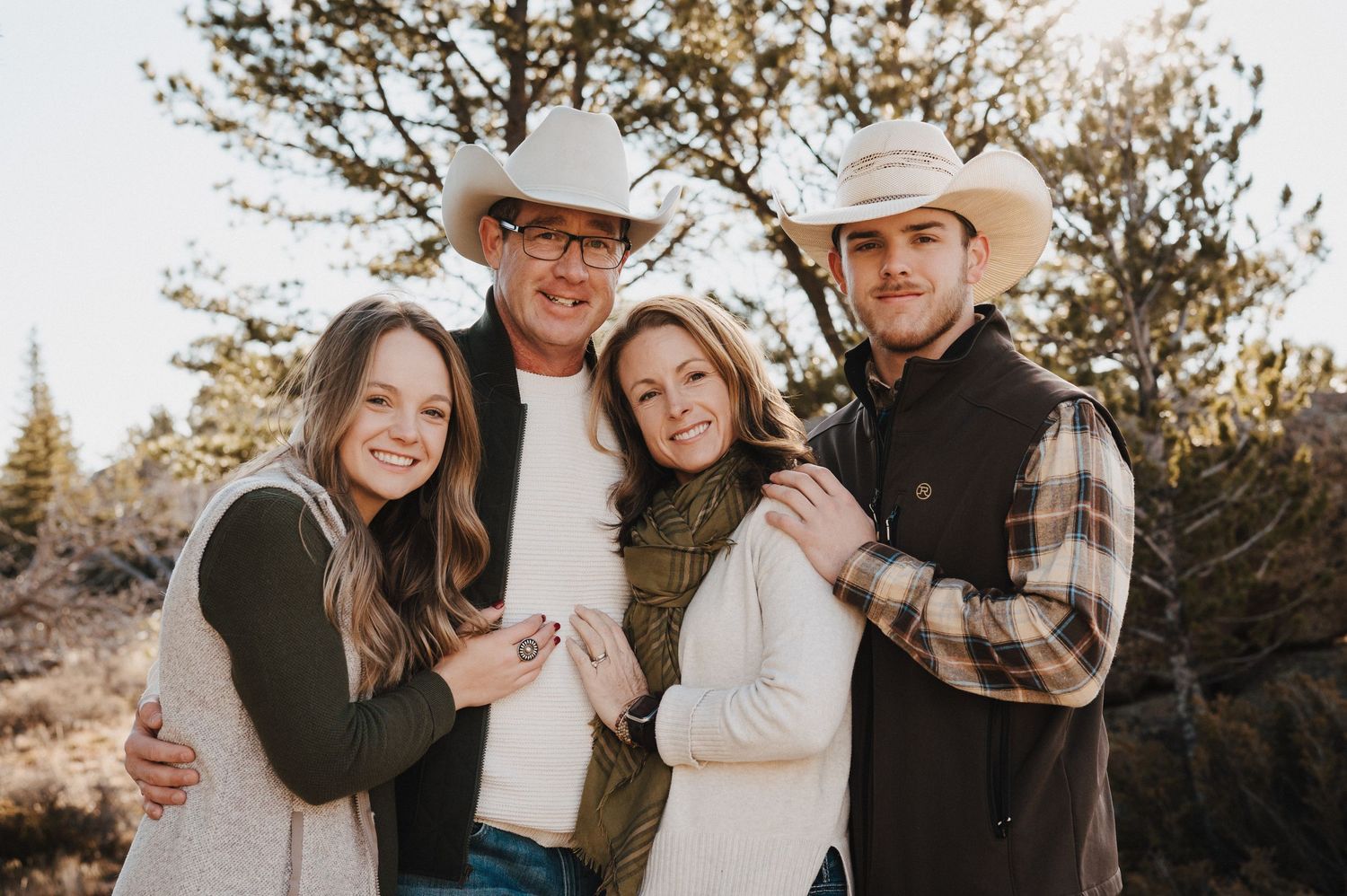 A group of four people wearing casual winter attire pose together outdoors during golden hour in a natural setting.