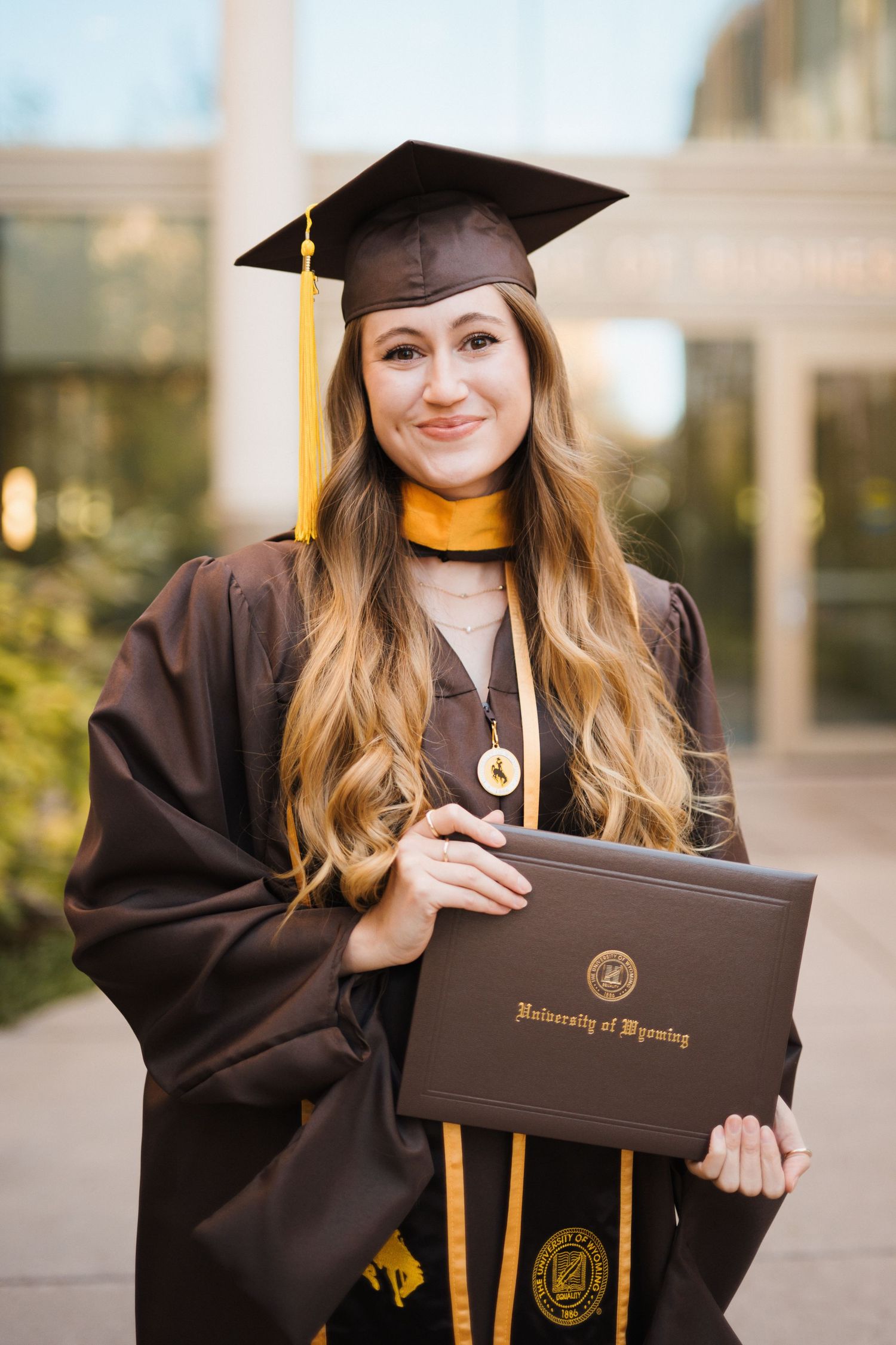 A college senior woman wearing graduation regalia holds a diploma in front of the University of Wyoming College of Business.