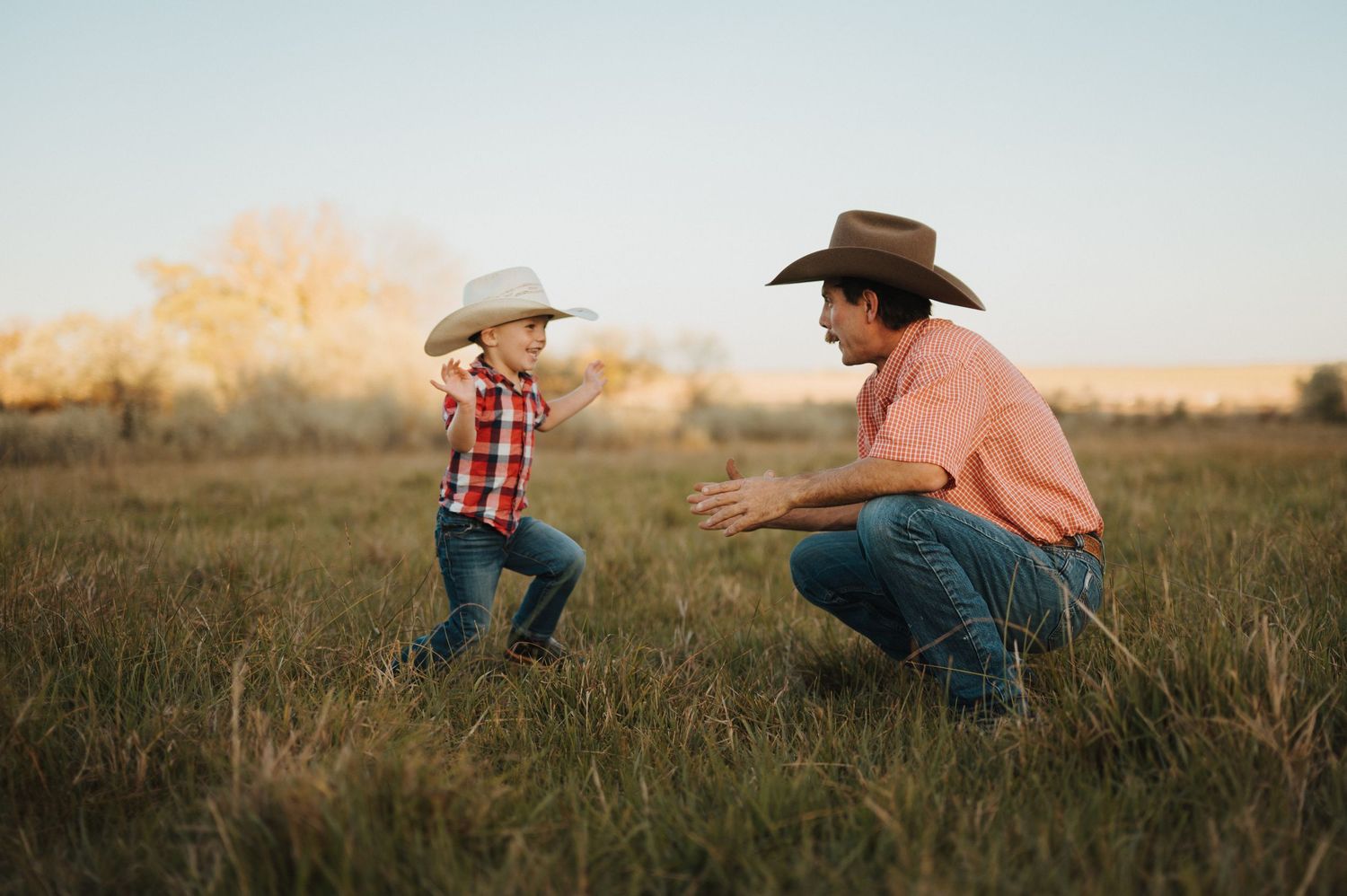 Two people in cowboy hats playfully chase each other through a golden field at sunset.
